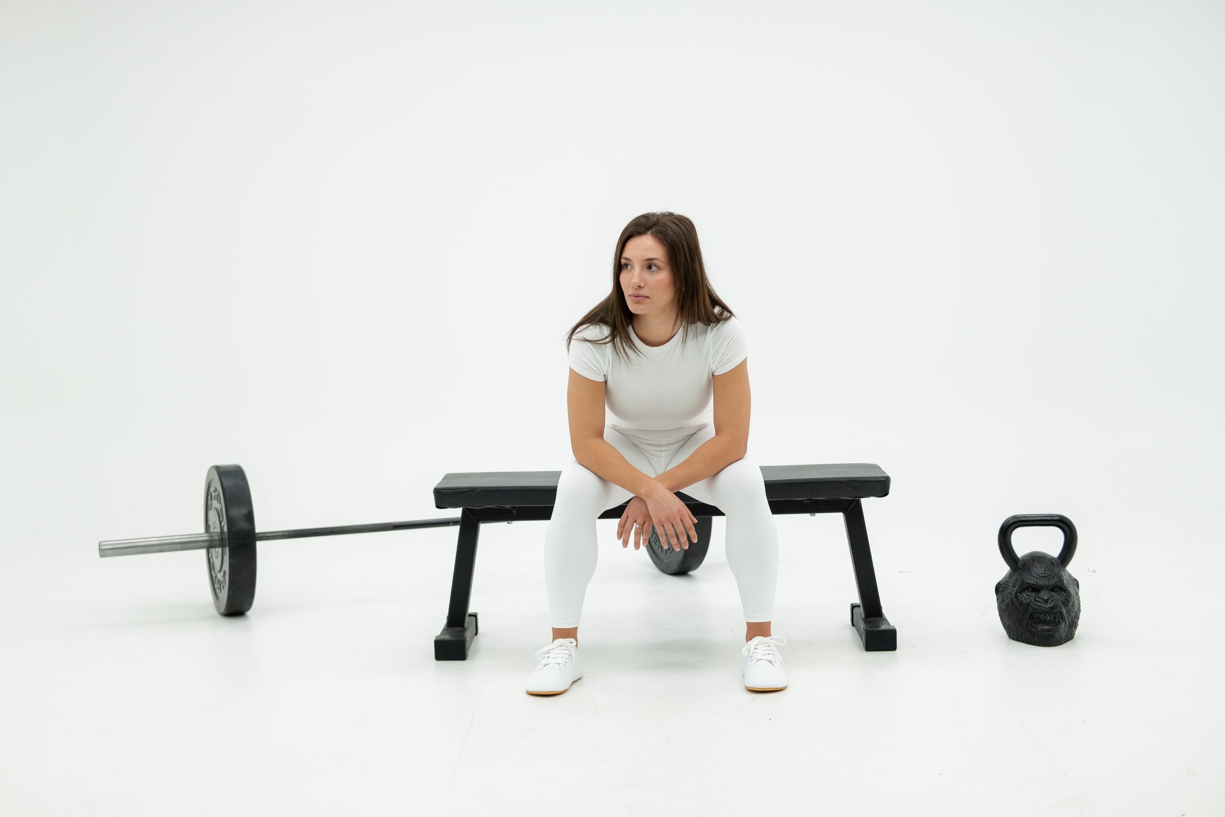 Woman sitting on bench with barbell and kettle bell against white backdrop.