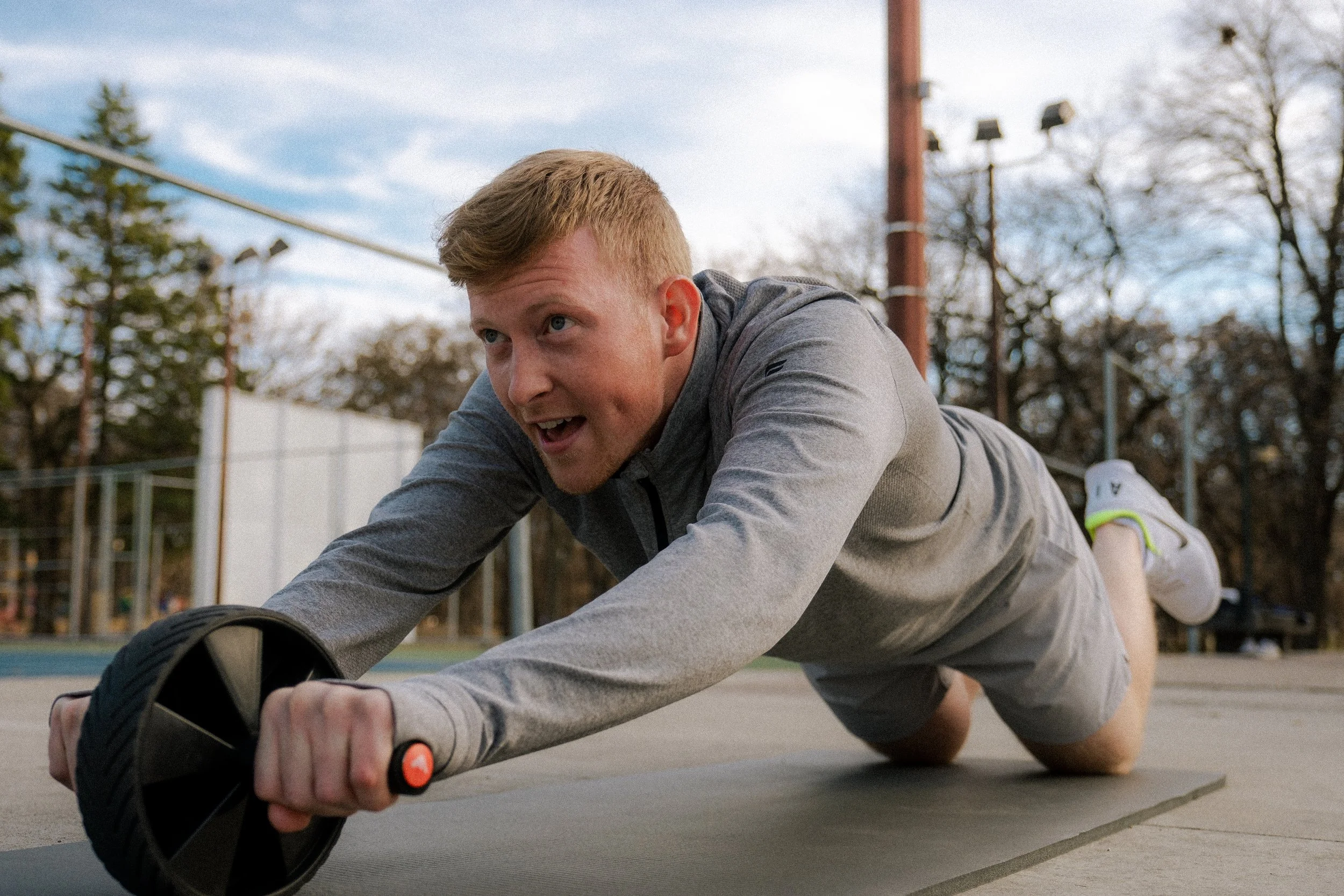 Man doing ab wheel workout outside, wearing grey long sleeve quarter zip jacket.