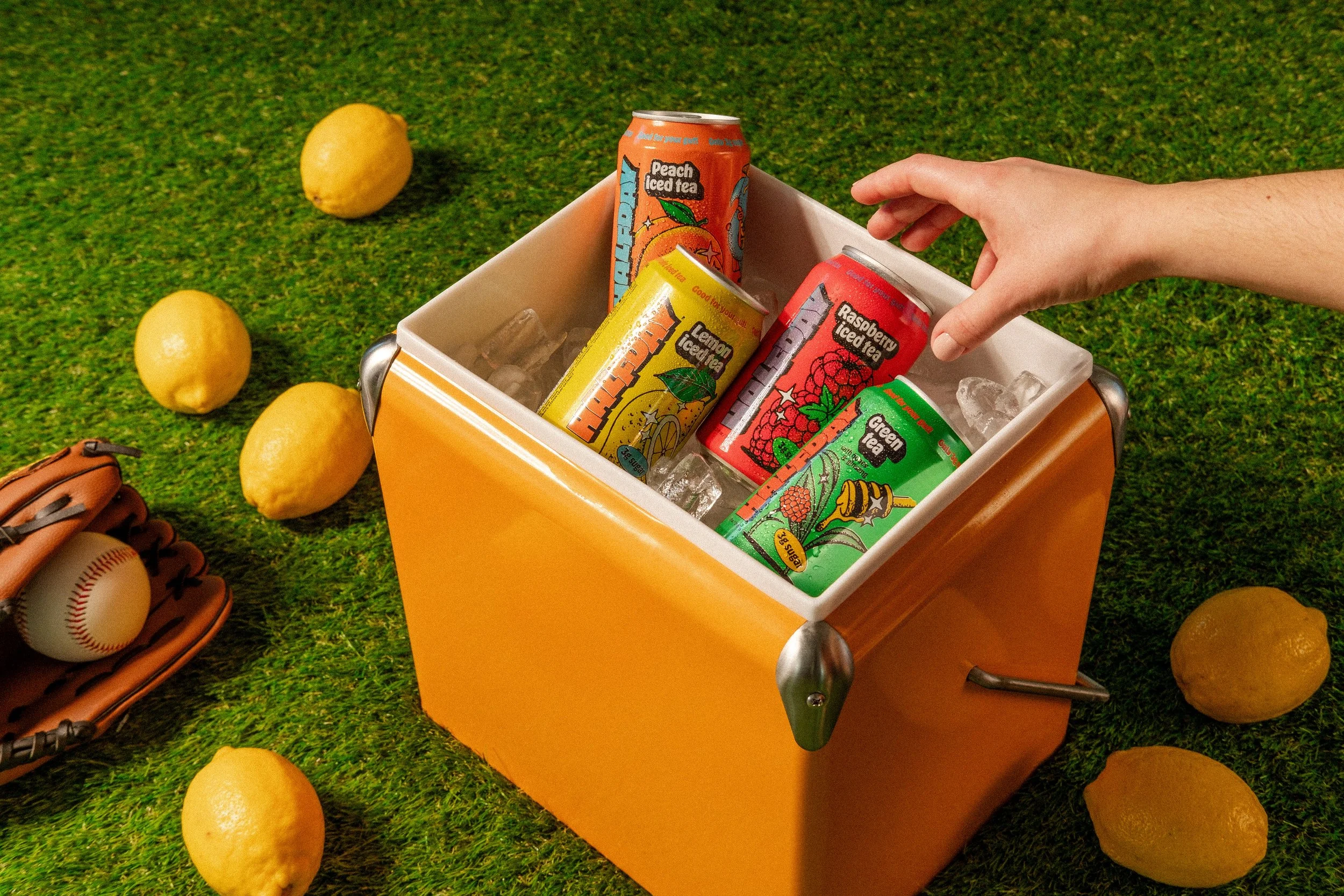 Female hand reaching into orange cooler with Halfday Iced Tea, and lemons surrounding.