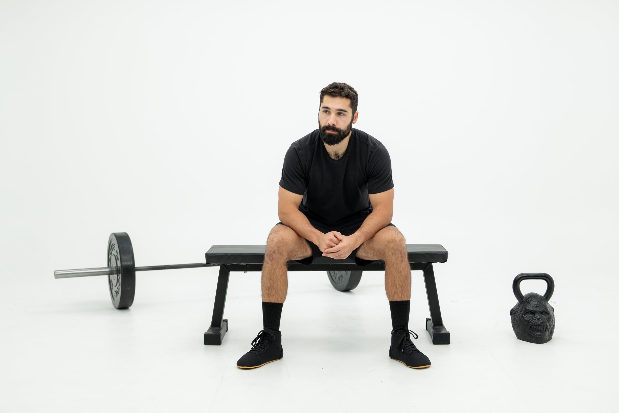 Man sitting on bench with barbell and kettle bell against white backdrop.