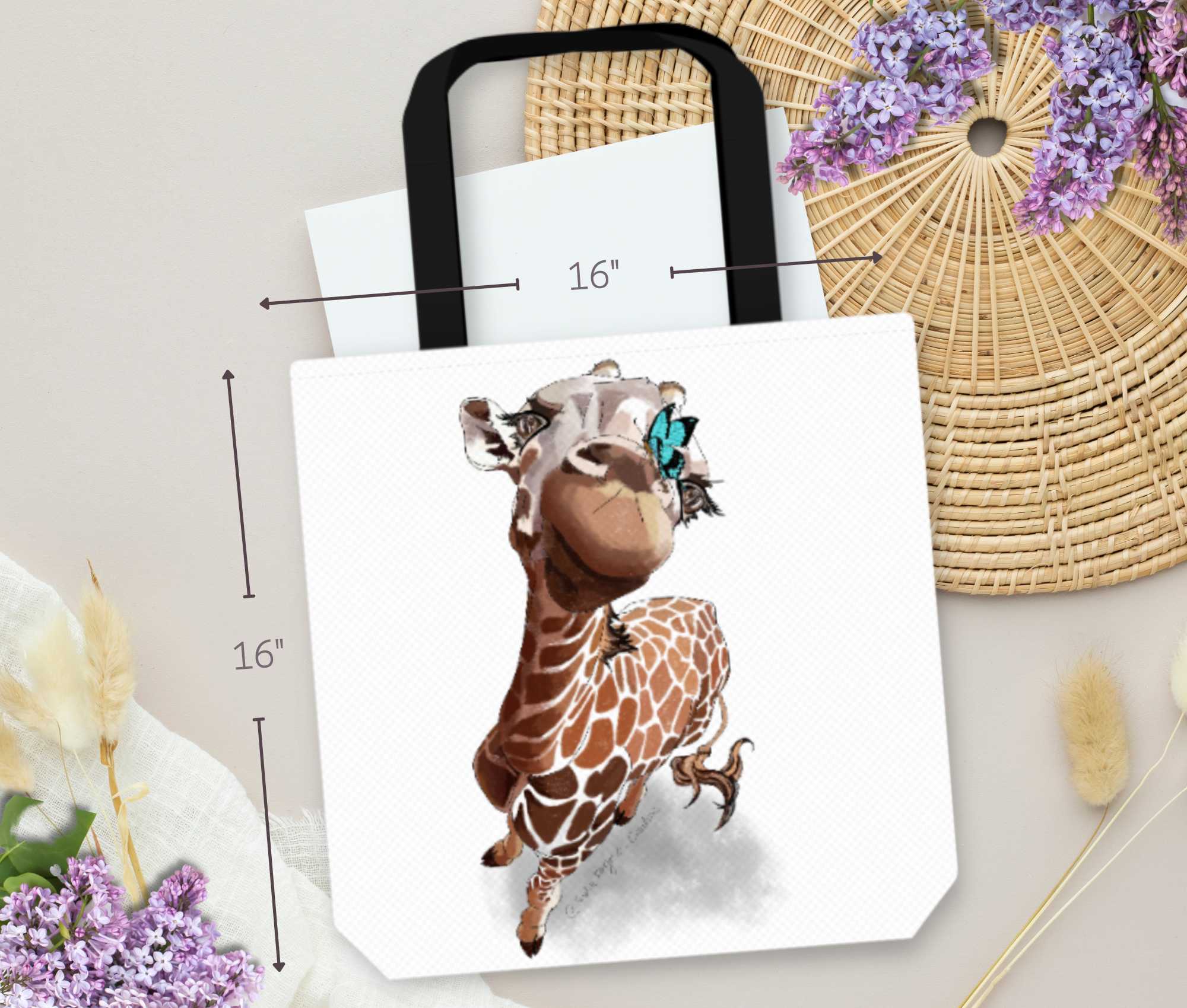 A giraffe illustration on a 16-inch black handled tote bag, resting on a round woven straw mat, purple flowers, and dried white grasses on a light background.