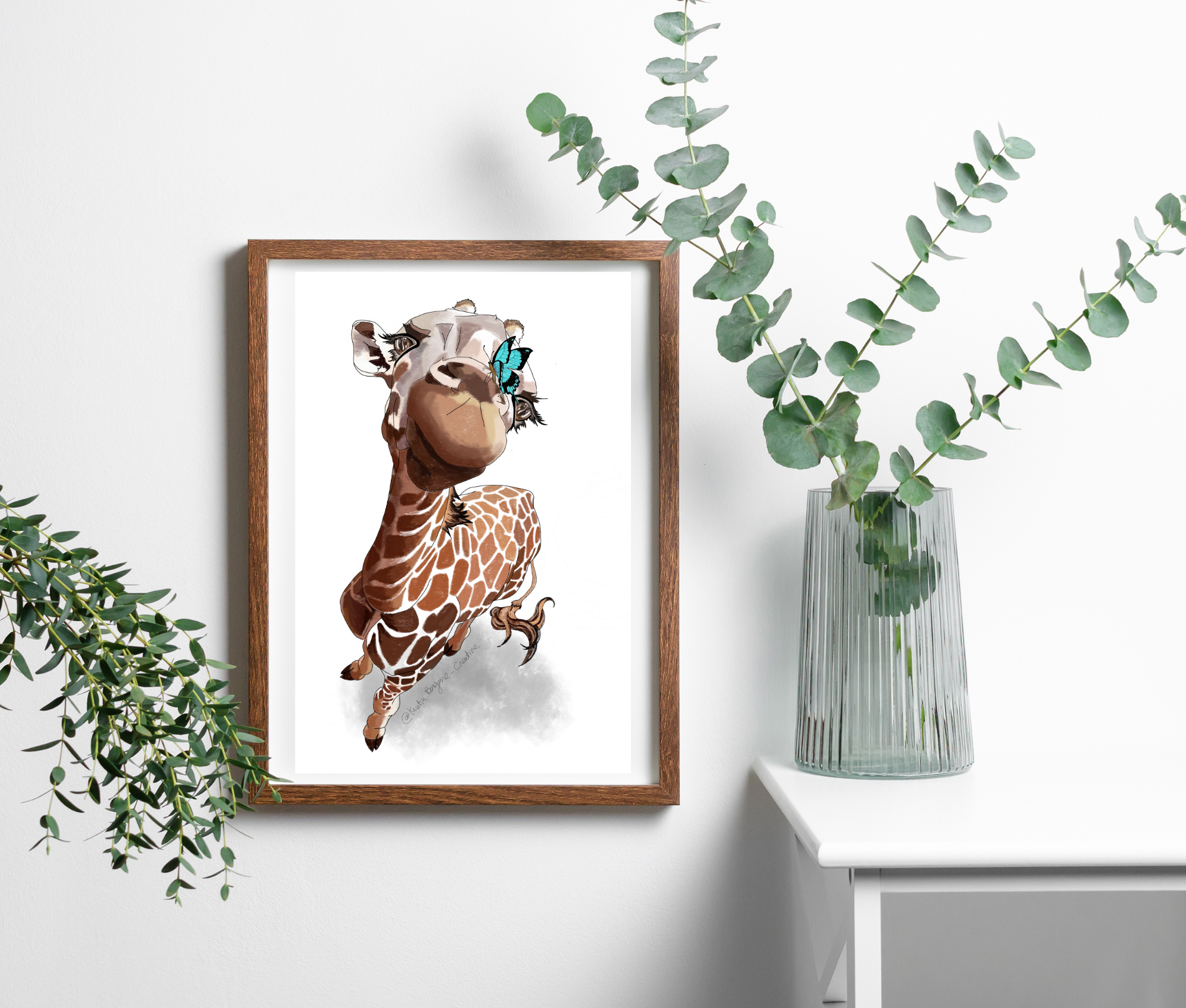 A framed illustration of a cartoon giraffe with a butterfly on its face, hanging on a white wall next to a glass vase with green eucalyptus branches.
