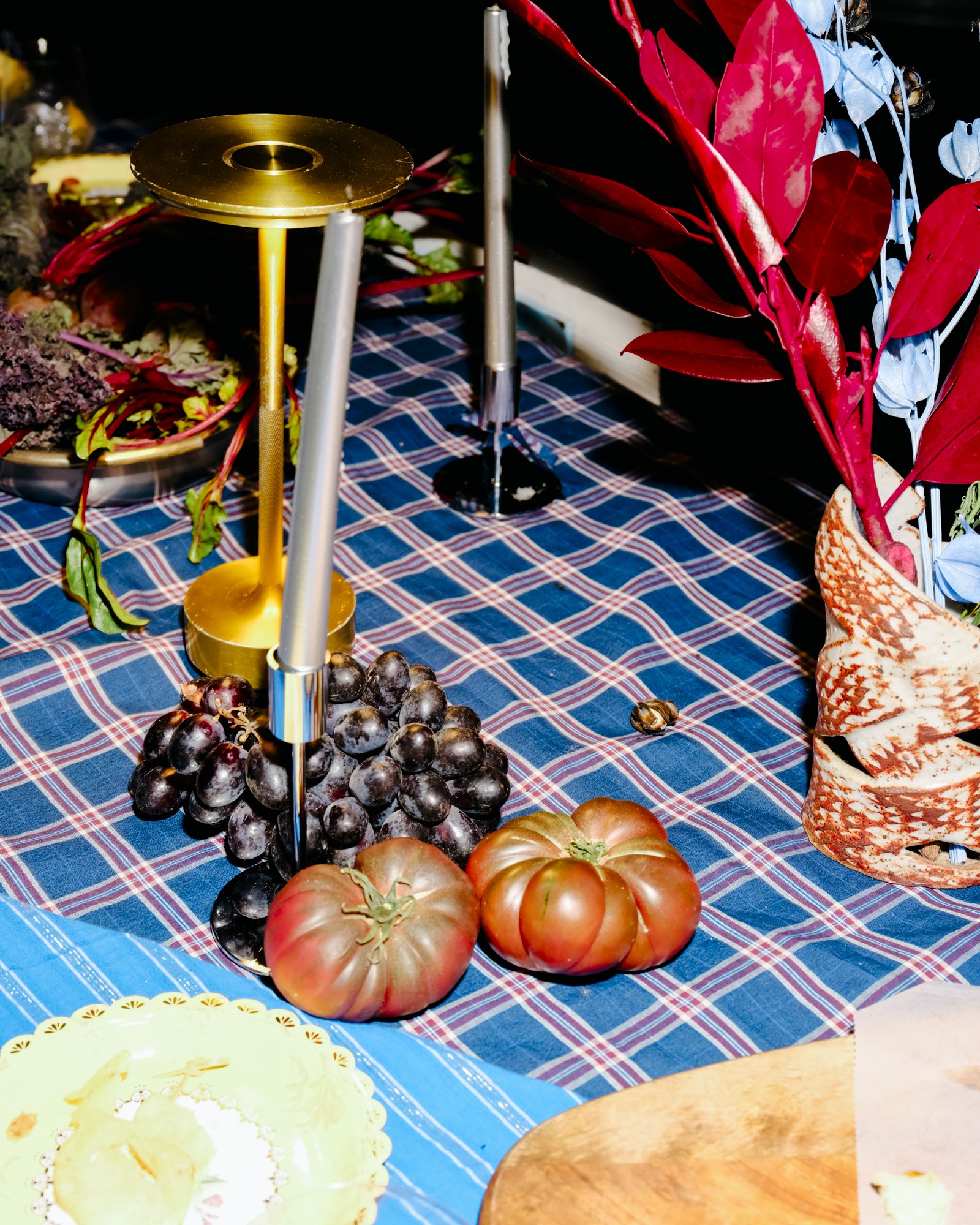 Grapes and tomatoes on a checkered tablecloth with candles and flowers in the background.