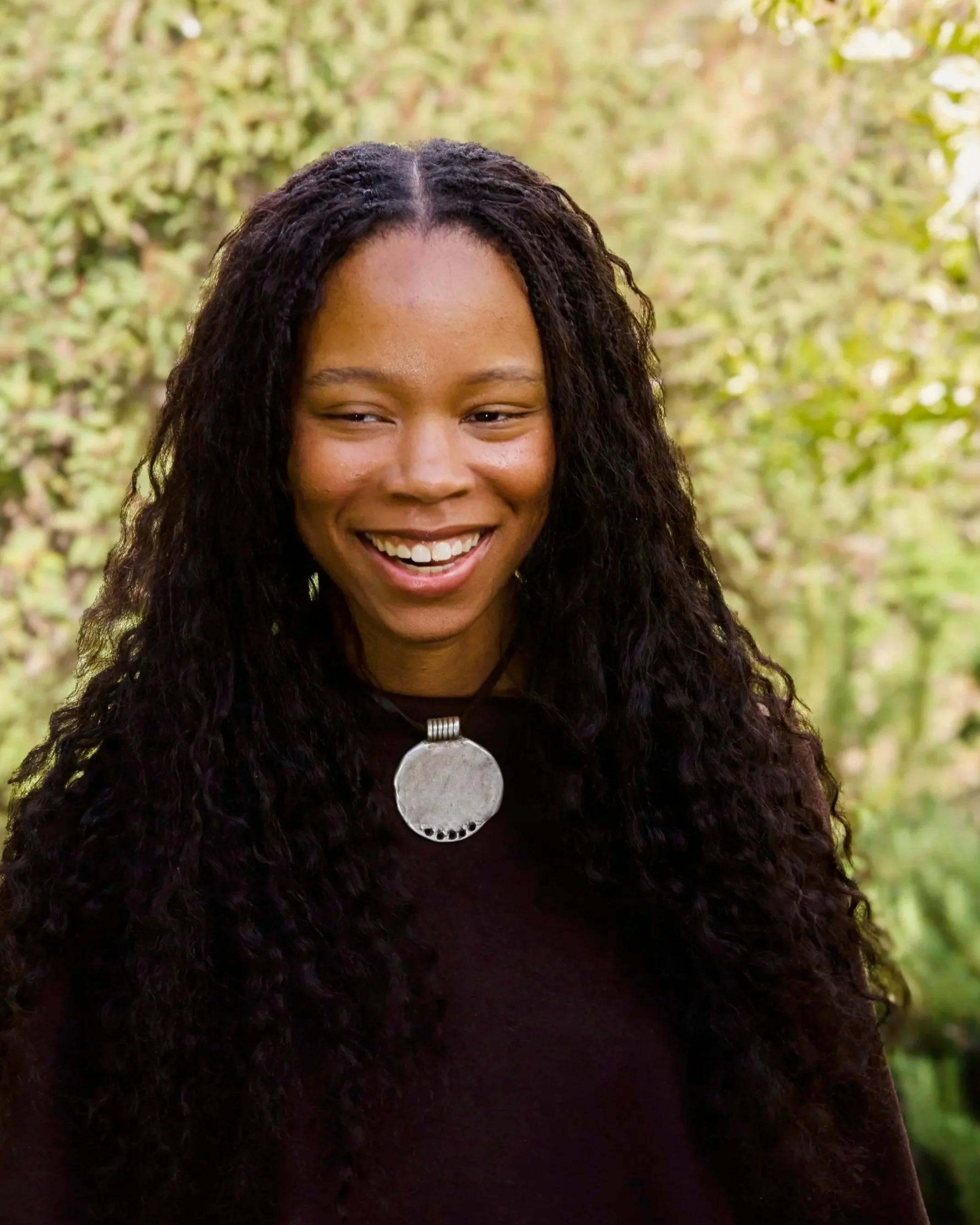 A woman with long, curly black hair smiling outdoors during daytime with green and yellow foliage in the background.