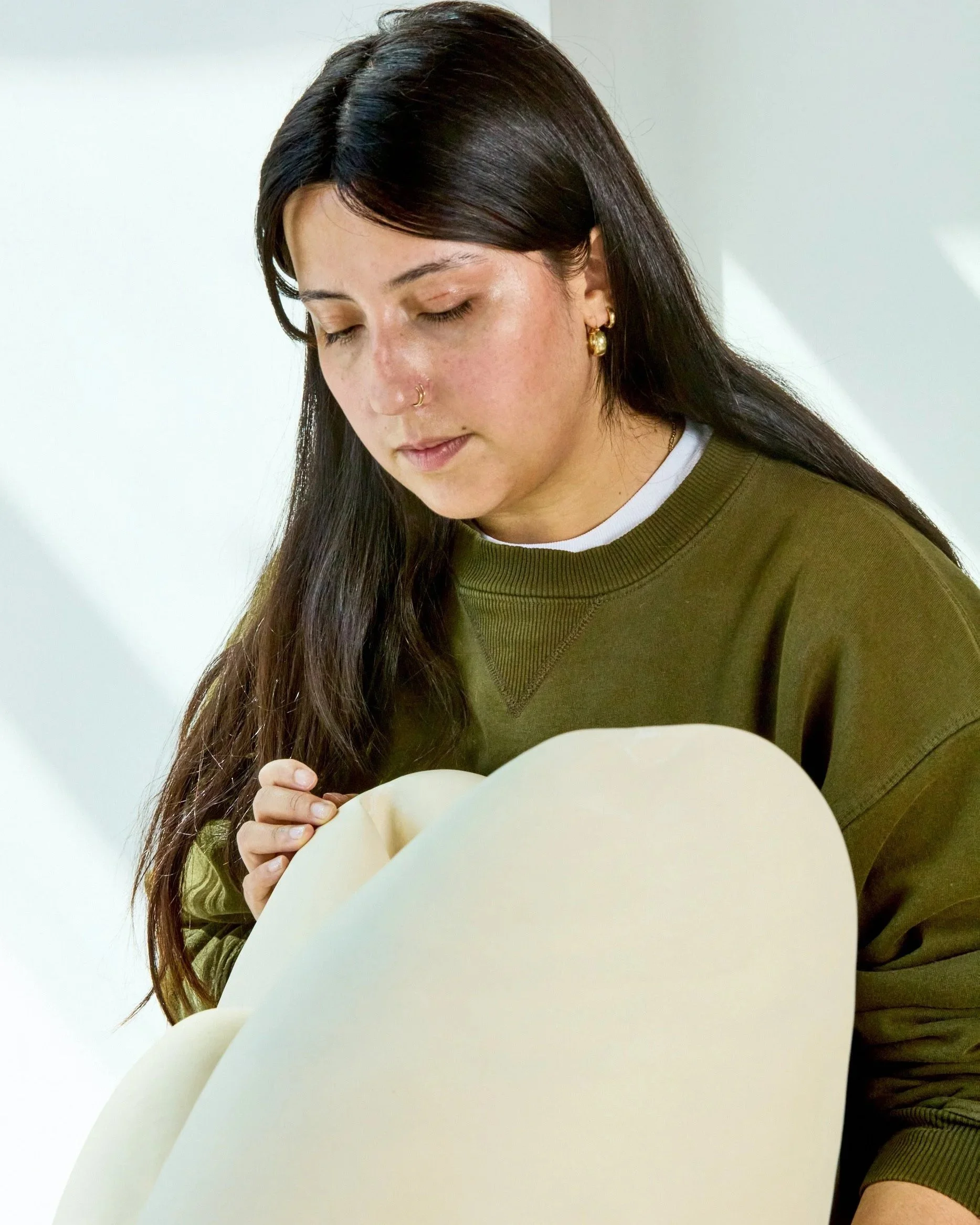 Young woman with long dark hair, gold earrings, and a nose ring, wearing an olive green sweatshirt, looking down at a large sheet of beige paper.