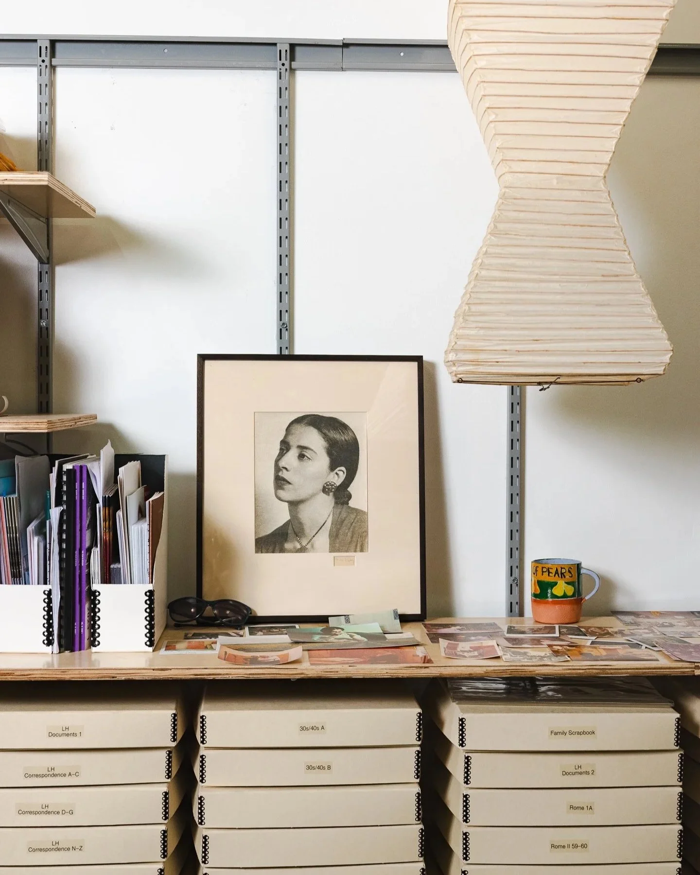 A cluttered office or storage space with a framed black-and-white portrait of a woman on the desk, next to a pair of sunglasses, and surrounded by file organizers, photographs, and a mug with the word 'pears' on it. A large, beige, hourglass-shaped p