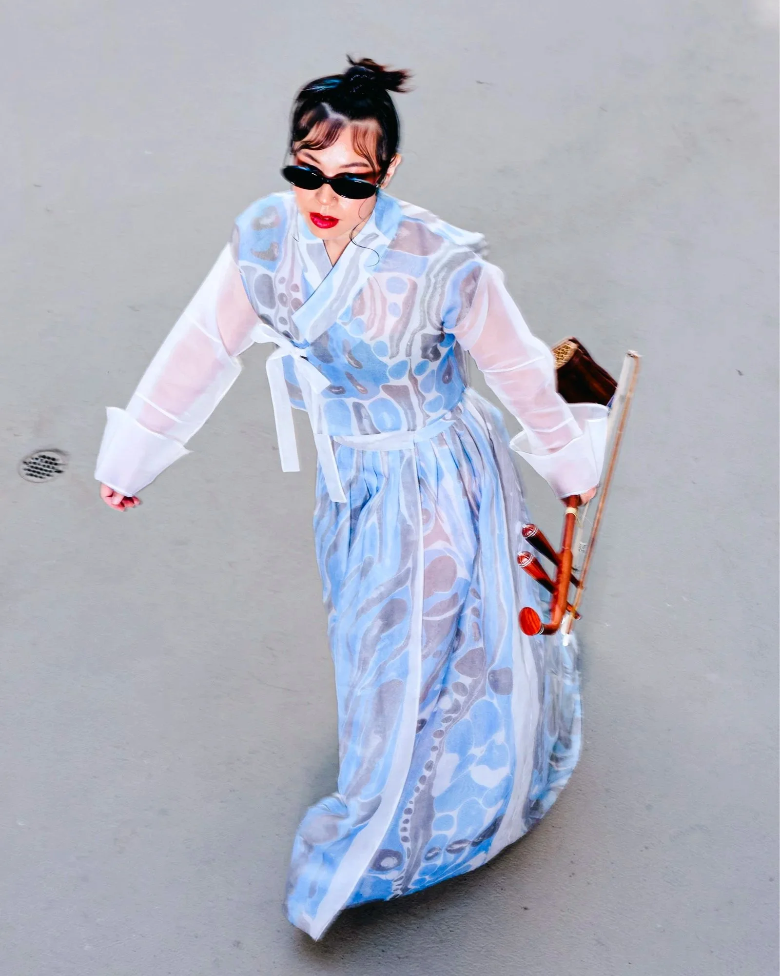 A woman dressed in a sheer, patterned blue and white kimono, wearing sunglasses with red lipstick, walking on gray pavement while carrying a traditional stringed instrument.