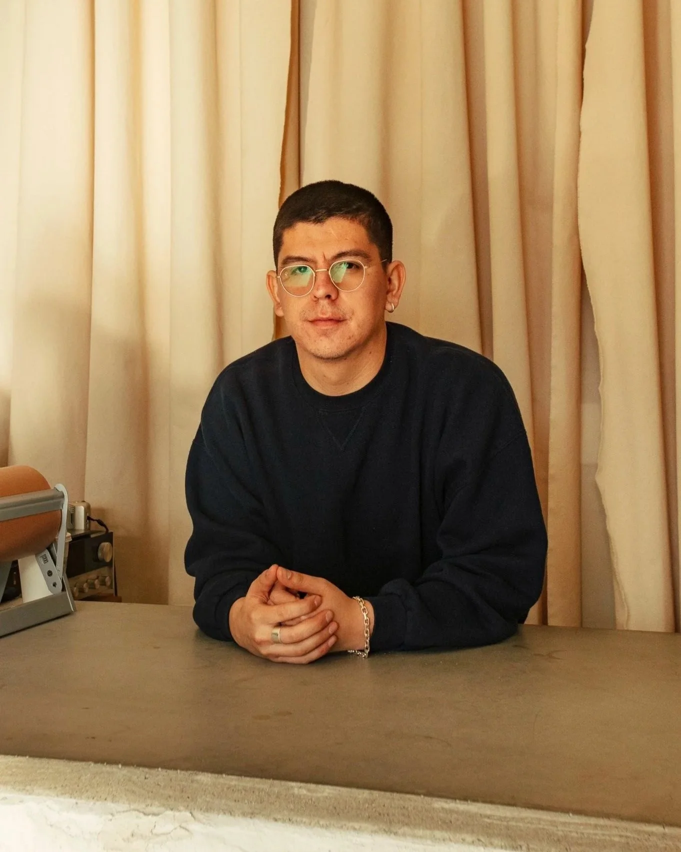 A man with short dark hair, glasses, and earrings sitting at a desk in front of beige curtains, wearing a black sweatshirt.