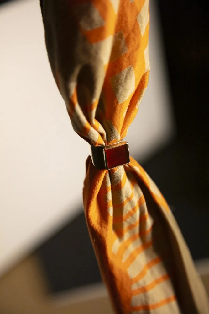 Close-up of a fabric curtain with an orange, beige, and brown pattern, gathered and secured with a black square clasp.