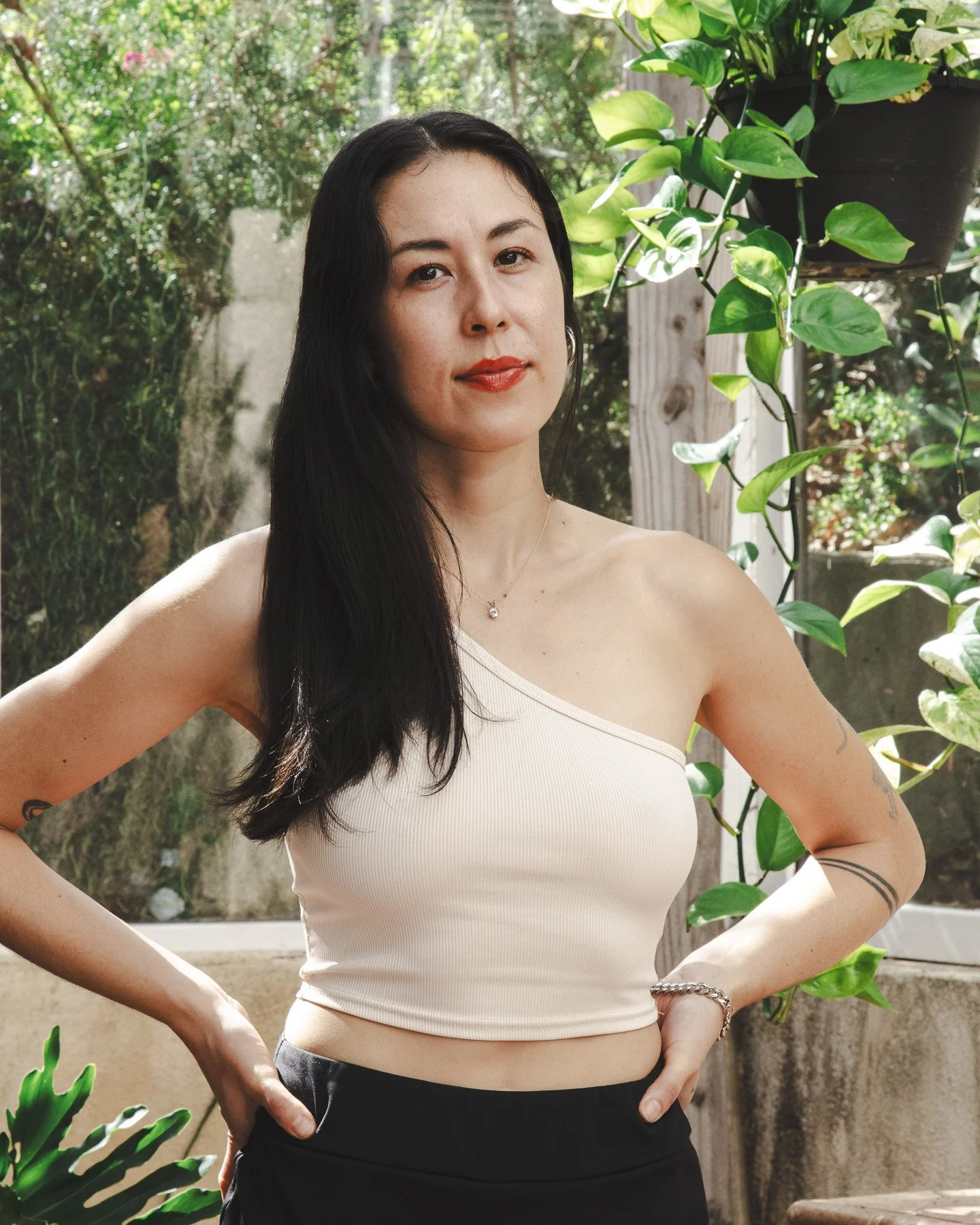 A woman with long black hair, wearing a cream-colored, one-shoulder crop top and black pants, standing outdoors in a garden with lush green plants and trees.