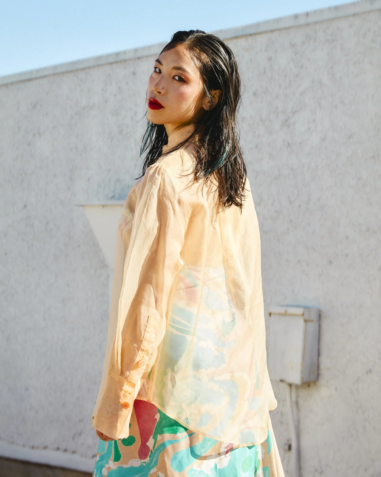 A woman with black, slightly wavy hair and bold red lipstick standing outdoors against a white wall, wearing a sheer, peach-colored blouse and a floral skirt with pastel shades, looking over her shoulder.