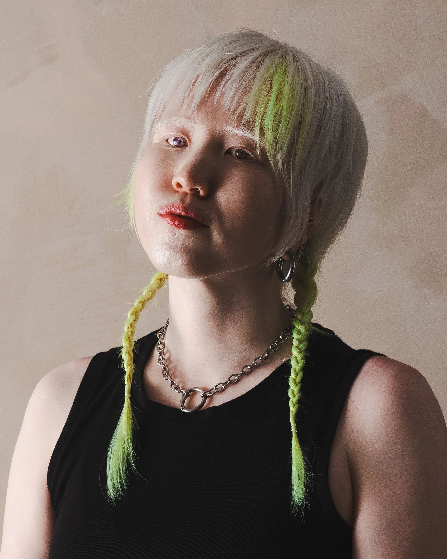 A young person with platinum blonde hair with neon green accents in two braids, wearing a black sleeveless top, silver chain necklaces, and hoop earrings, against a beige background.