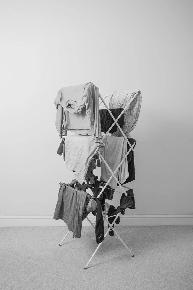 A foldable drying rack filled with various hanging clothes in a room with a plain wall and carpeted floor.