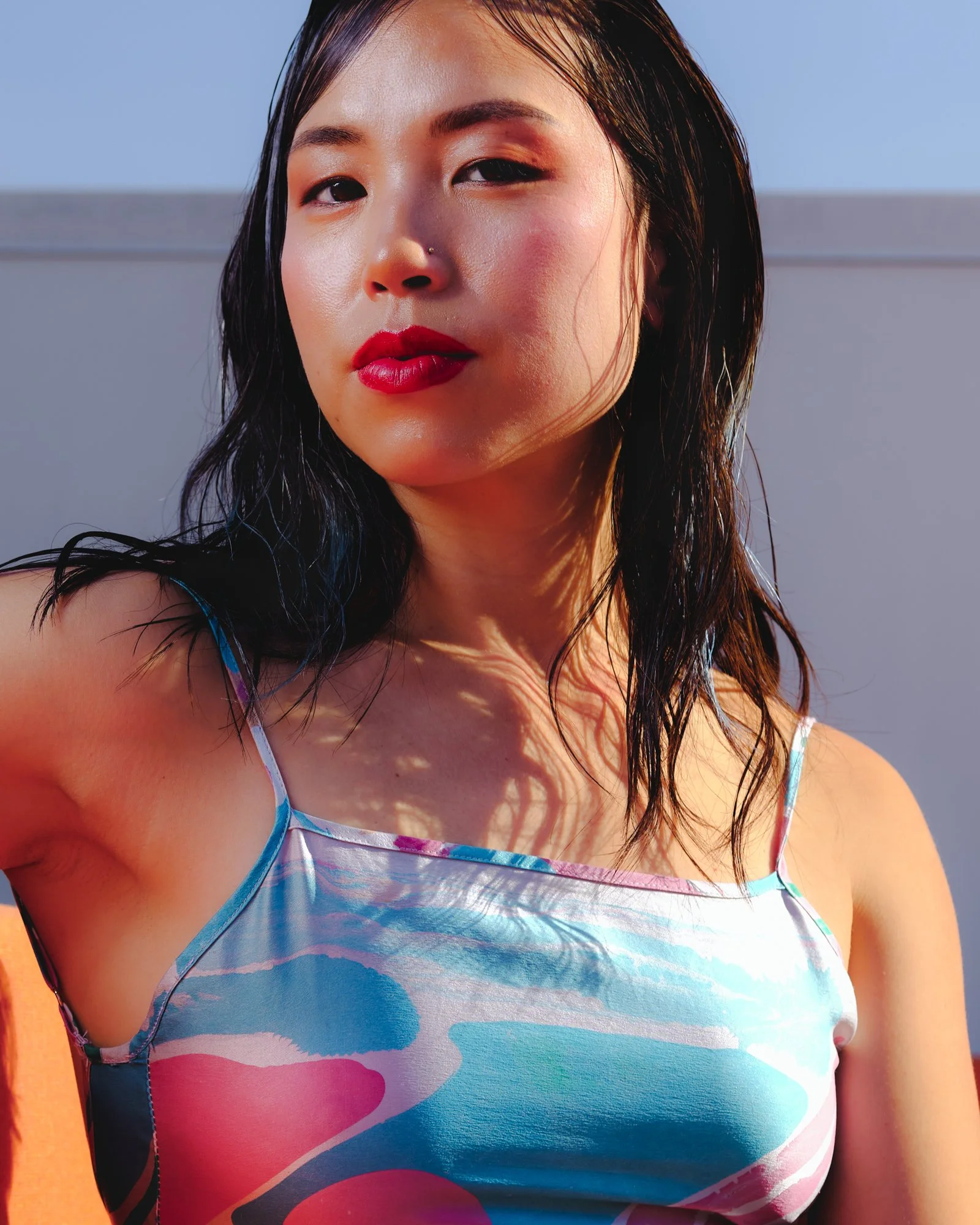 A woman with wavy dark hair wearing a colorful, tie-dye spaghetti strap top, with sunlight casting shadows on her face and shoulder, against a background with clear blue sky and a gray horizontal surface.