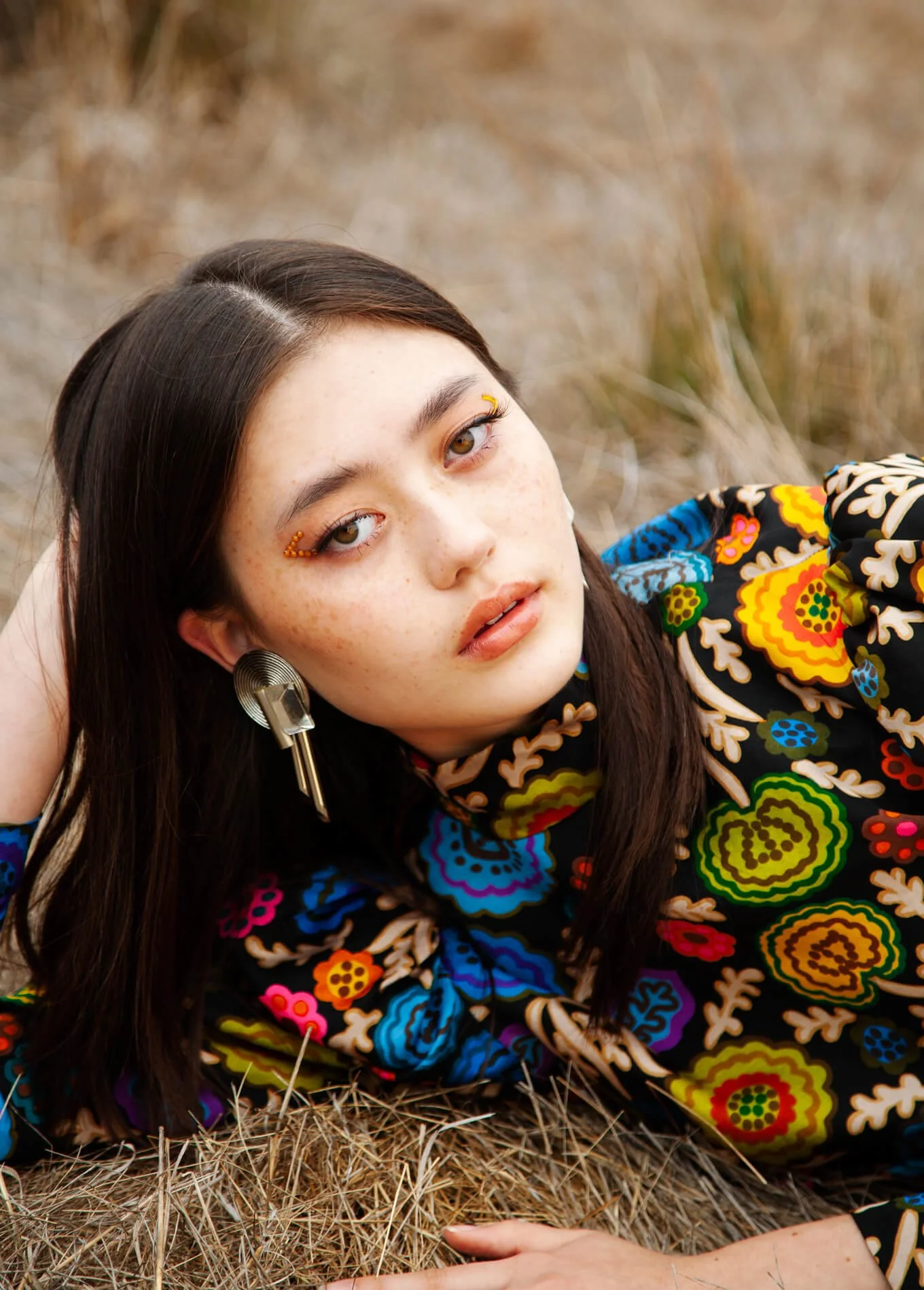 Young woman with brown hair lying on dry grass, wearing colorful floral patterned dress, with distinctive eyelash decorations and a large metallic earring.