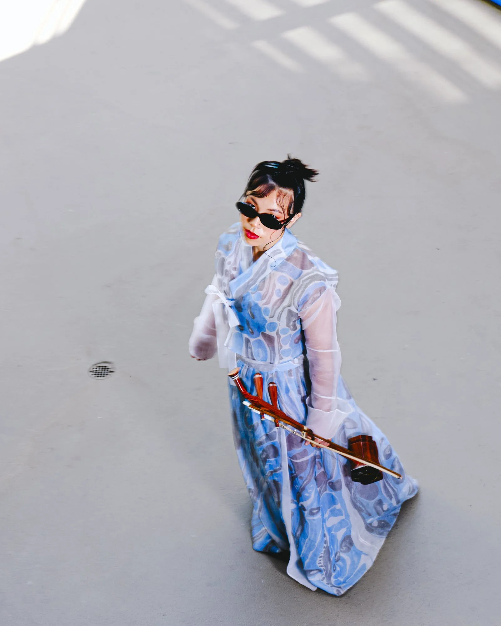 Woman with dark hair in a top knot, wearing black sunglasses and red lipstick, dressed in a flowing blue and white patterned dress, standing on a concrete surface, holding a long wooden object with a black bowl attached at one end.