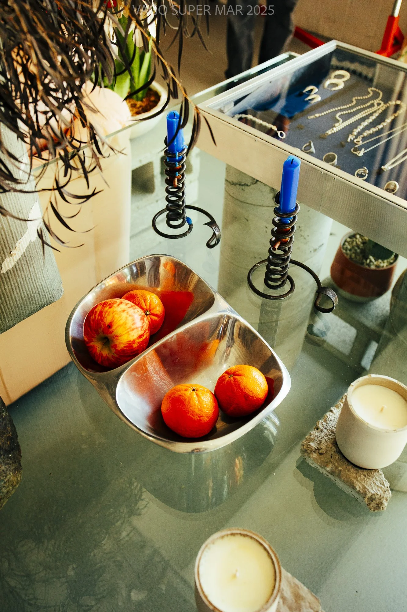 A glass table with a metallic divided bowl containing two apples and two oranges. There are candles placed on stone slabs and a decorative display of jewelry in a glass case in the background.