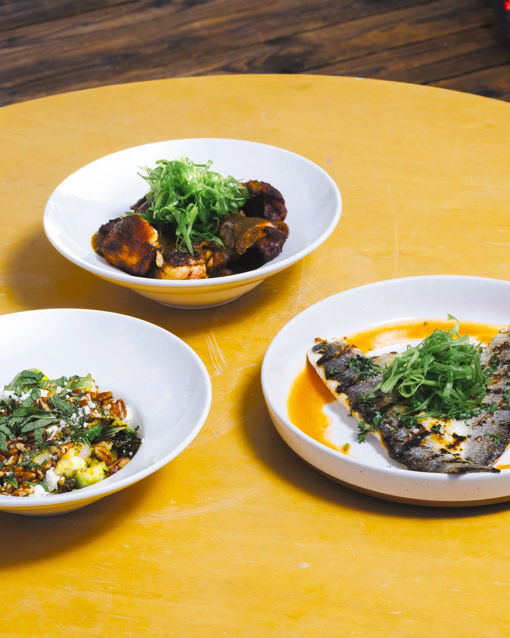Three bowls of Asian-inspired dishes on a yellow wooden table, featuring grilled fish garnished with green onions, a bowl of beef with greens, and a bowl of vegetables with herbs.