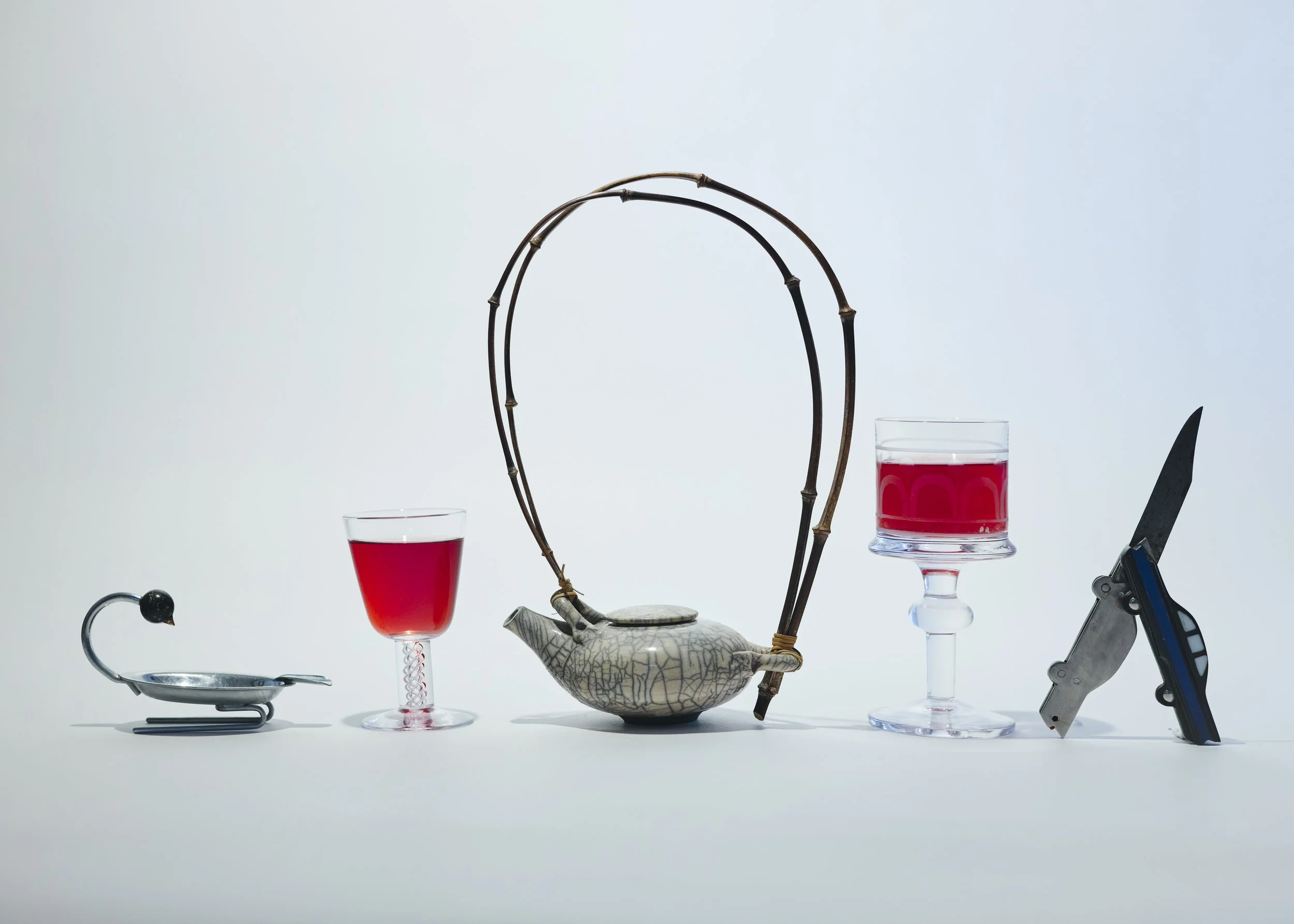 Scene of various objects related to tea and cutlery arranged on a white surface against a light gradient background, including a small figurine, a glass with red liquid, a decorative teapot, a glass with red liquid on a stand, a knife, and a multitoo
