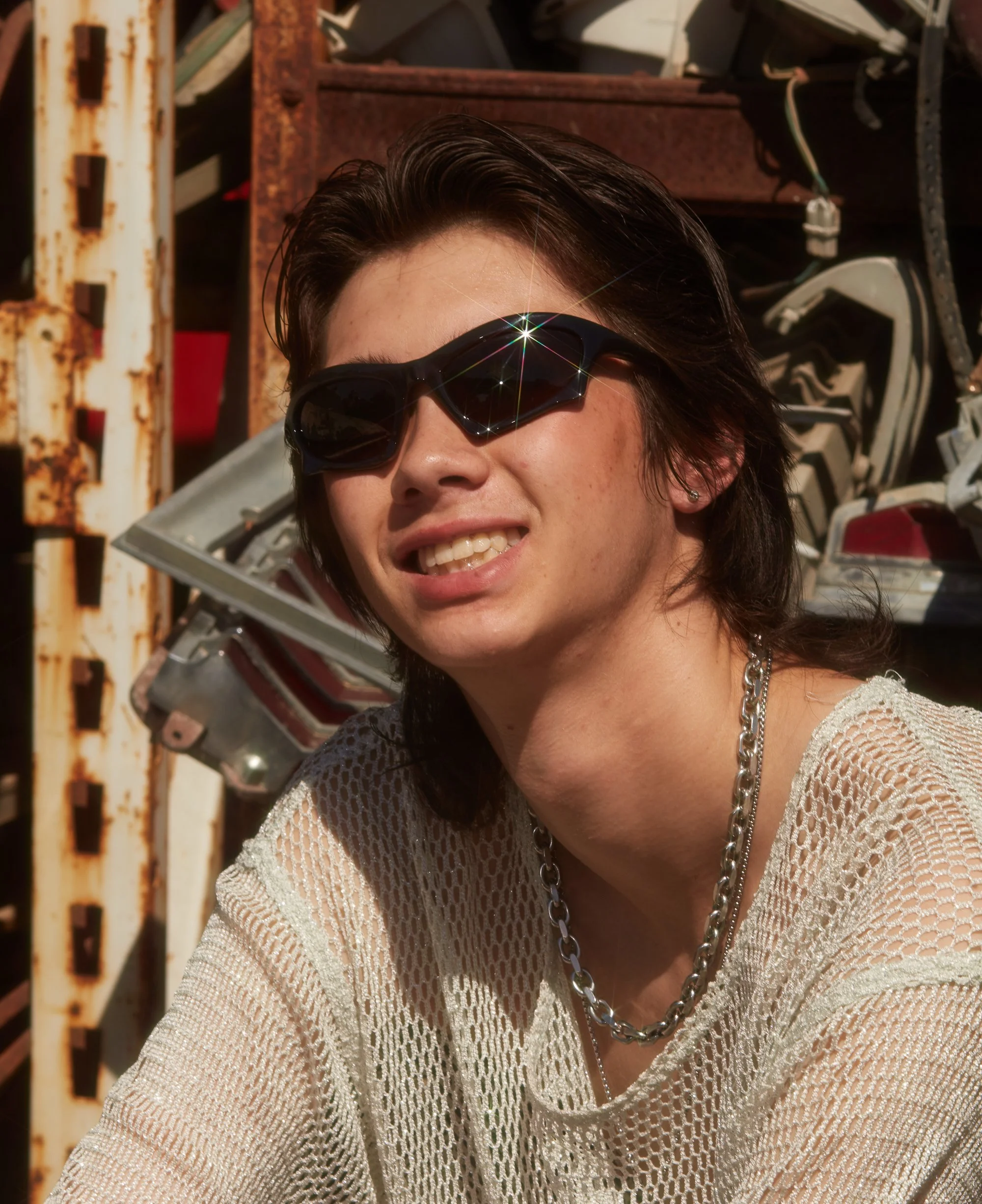 A young woman with dark hair wearing sunglasses, a mesh shirt, and a silver chain necklace, smiling outdoors with rusty metal and mechanical parts in the background.