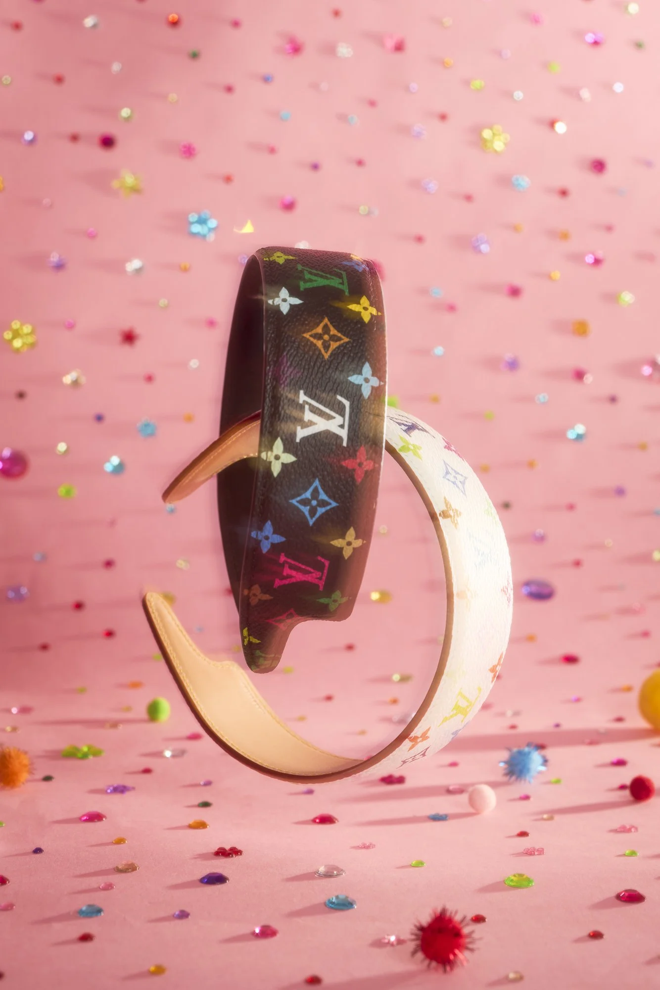 A colorful Louis Vuitton headband with multicolored logo patterns, floating against a pink background sprinkled with multicolored confetti and small decorative objects.