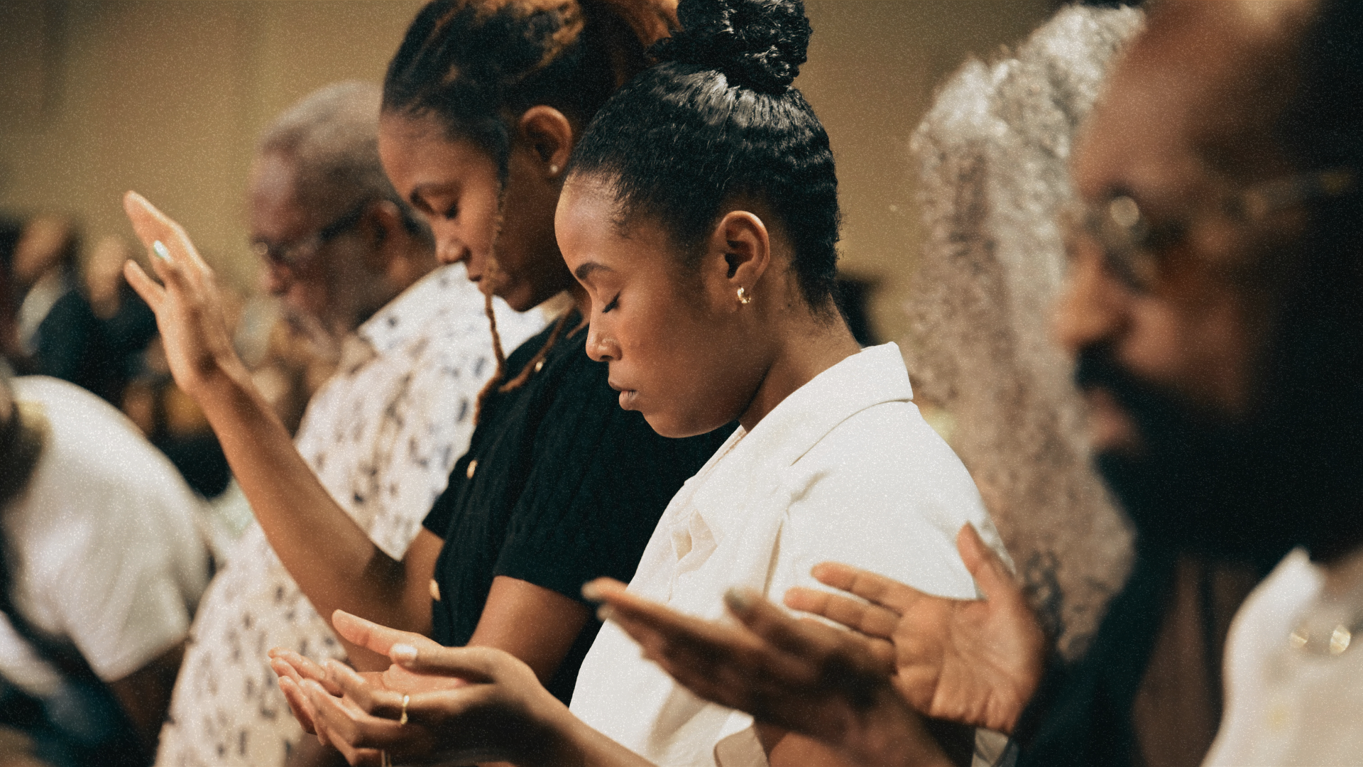 A man in a beige uniform with a patch on his arm is praying with his eyes closed and hands pressed together in front of his face, standing against a brick wall.