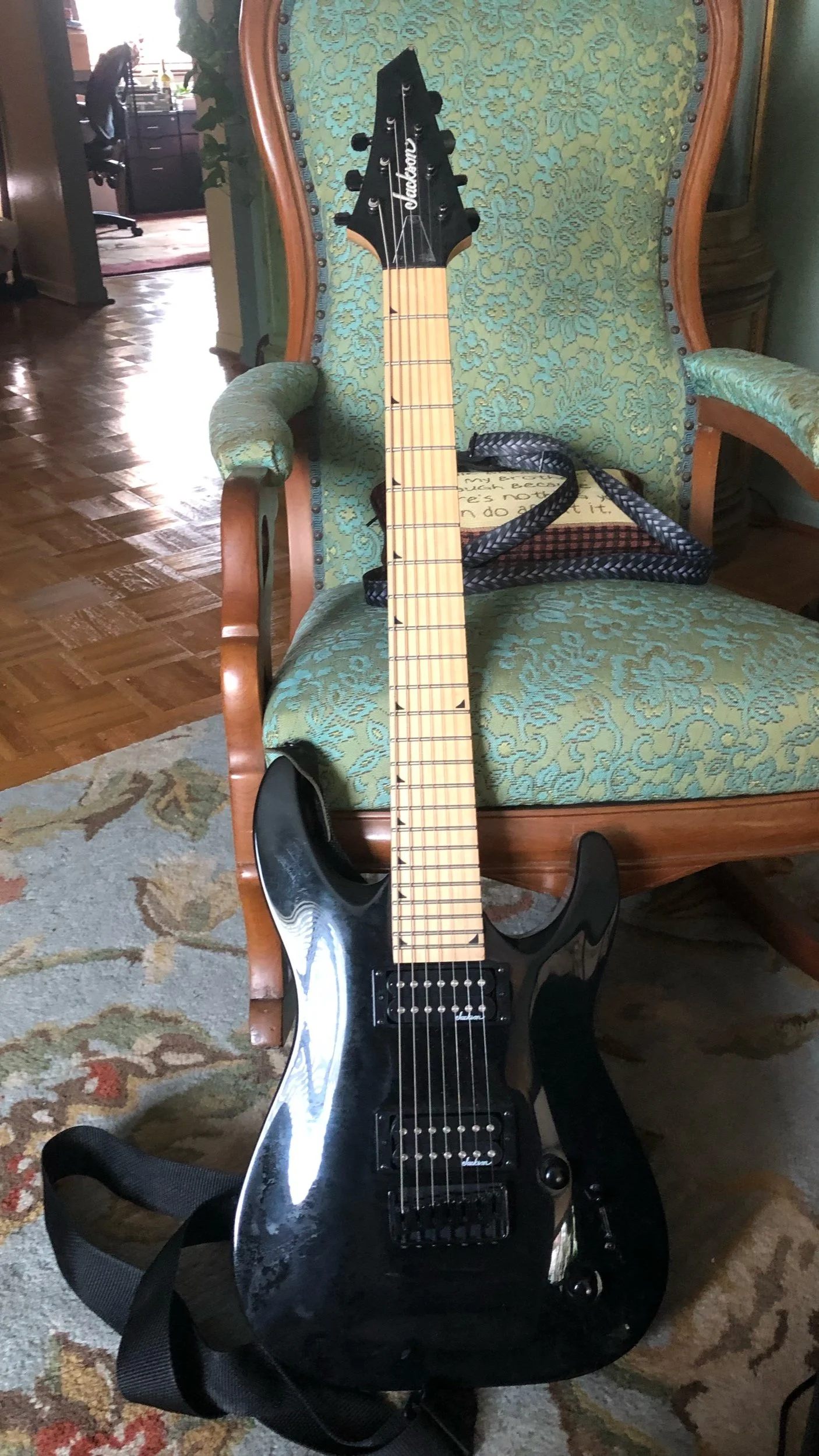 Black electric guitar with a light wood fretboard resting against a green upholstered armchair with wooden accents in a living room.