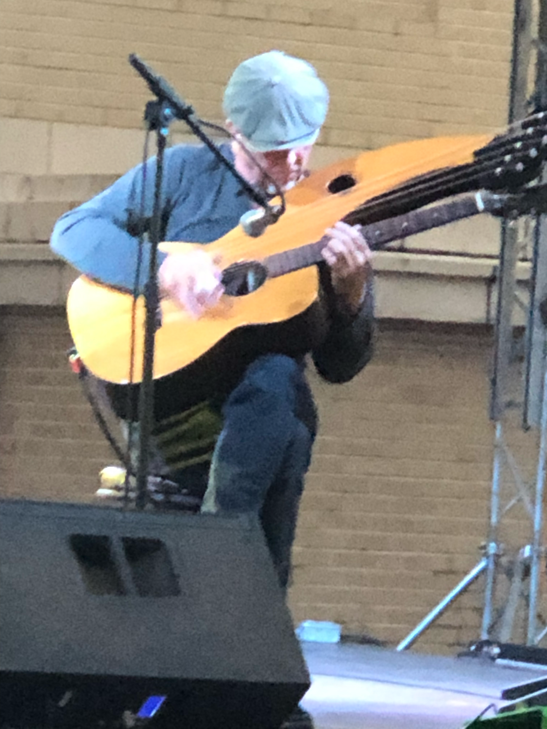 A person wearing a light blue cap and glasses is playing an acoustic guitar and singing into a microphone. They are standing in front of a wooden wall, with a music stand to their left.