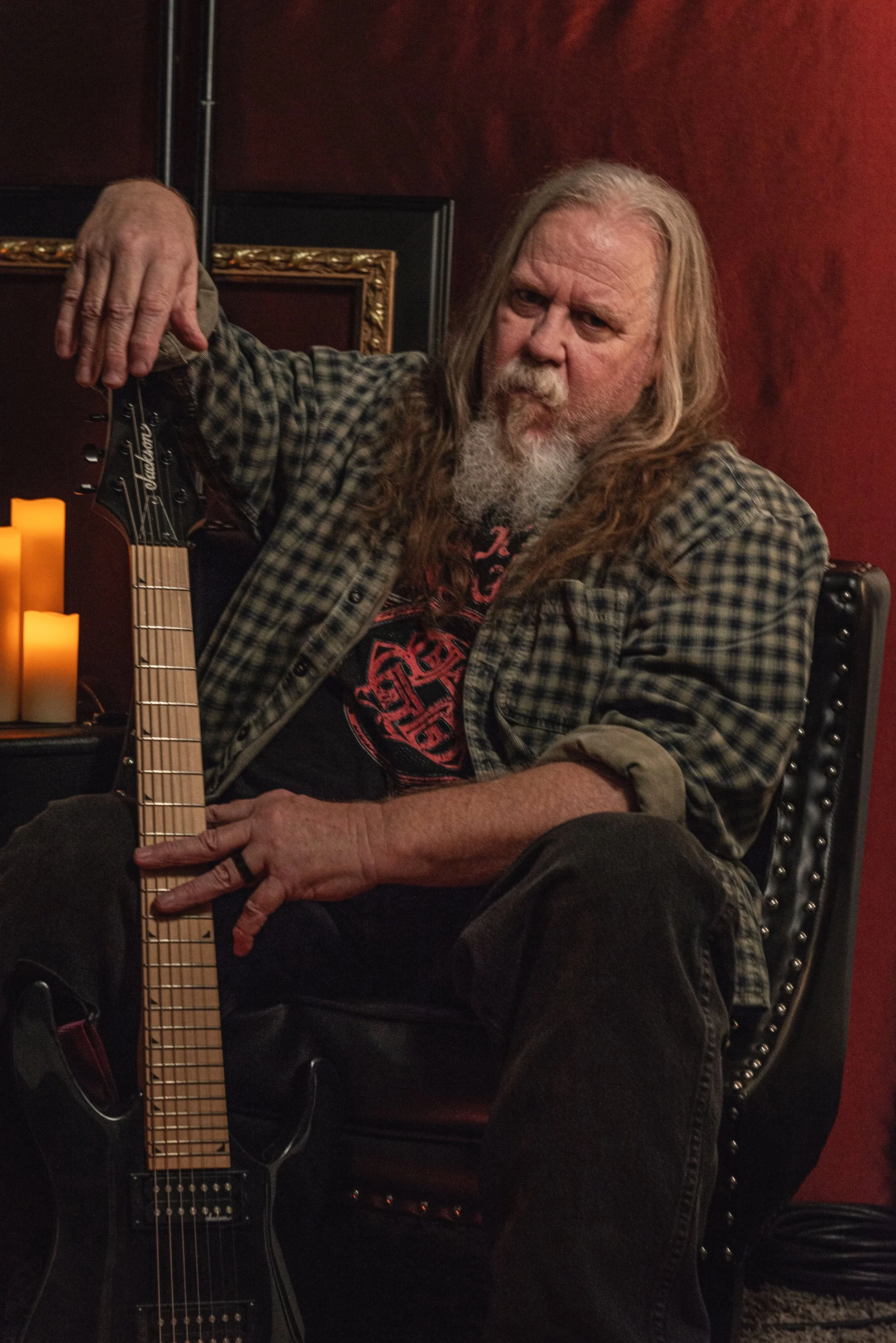 A photo of Kim Kimbo Groff with long gray hair and a beard sitting on a black leather chair, holding a black electric guitar, in a dimly lit room with a red wall and candles.