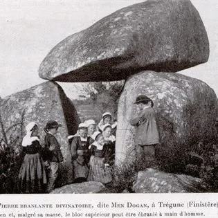 Historically dressed group of people, including women and children, gathered among large boulders in a natural outdoor setting.