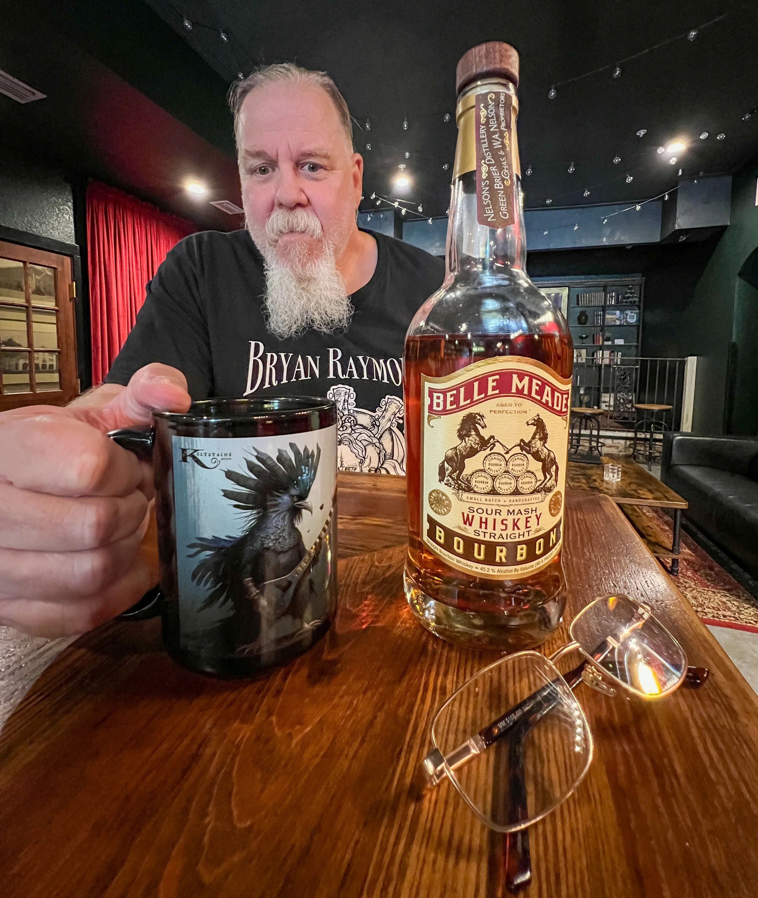 Kim Kimbo Groff with a beard sitting at a wooden table in a dimly lit bar, holding a black mug with a bird illustration, next to a bottle of Belle Meade bourbon whiskey, with glasses of whiskey and a pair of glasses on the table.