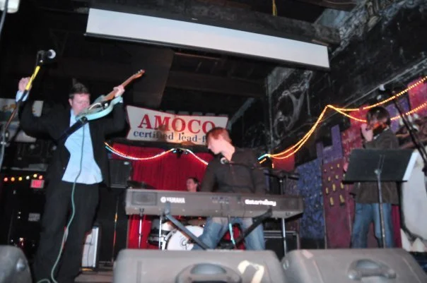 Band performing live on stage with a keyboard, microphone, and drums at a venue decorated with string lights and a banner that reads 'AMOCO certified lead-free'.