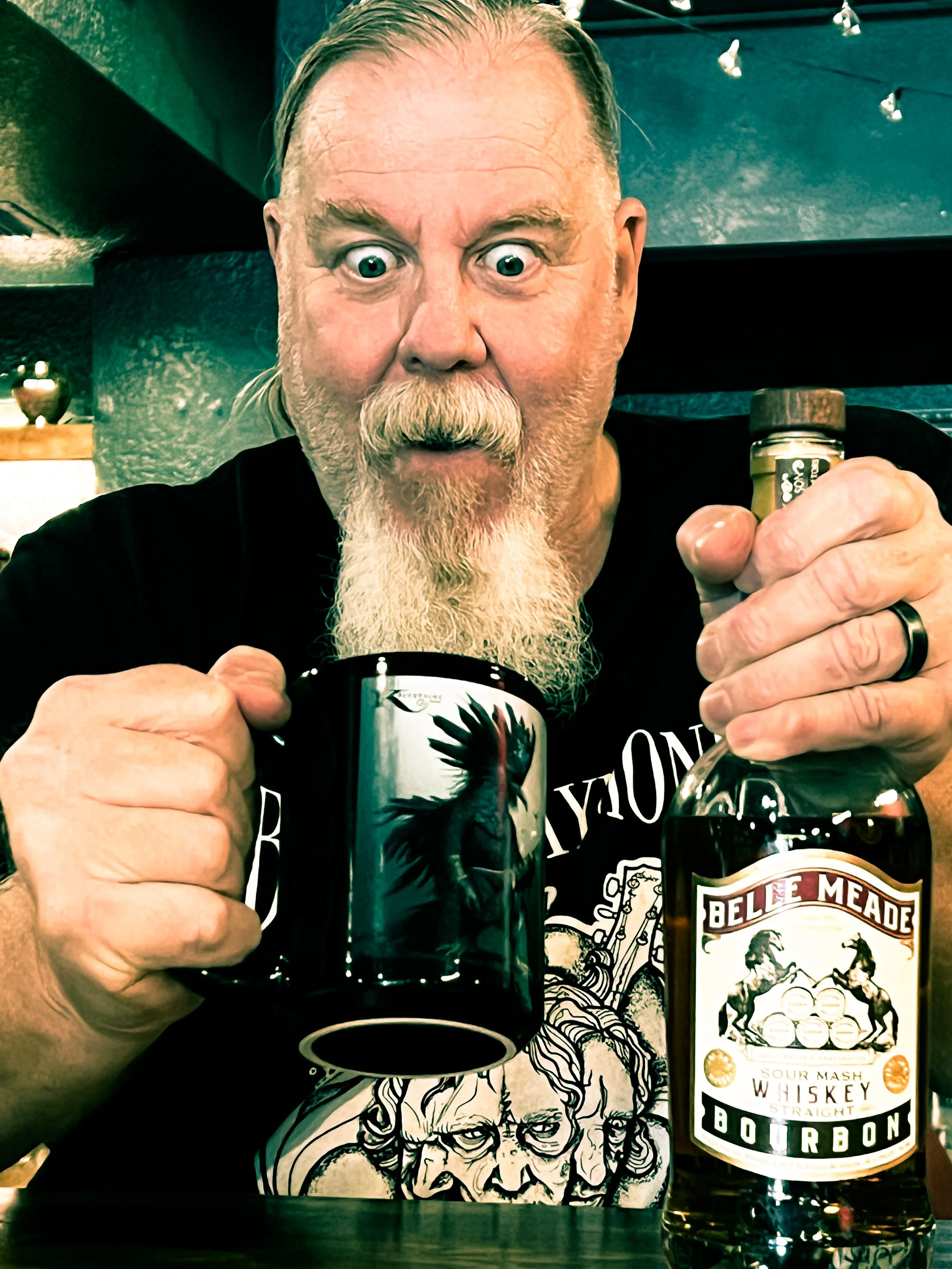 Kim Kimbo Groff with a surprised expression holding a black mug and a bottle of Belle Meade bourbon whiskey in a dimly lit room.