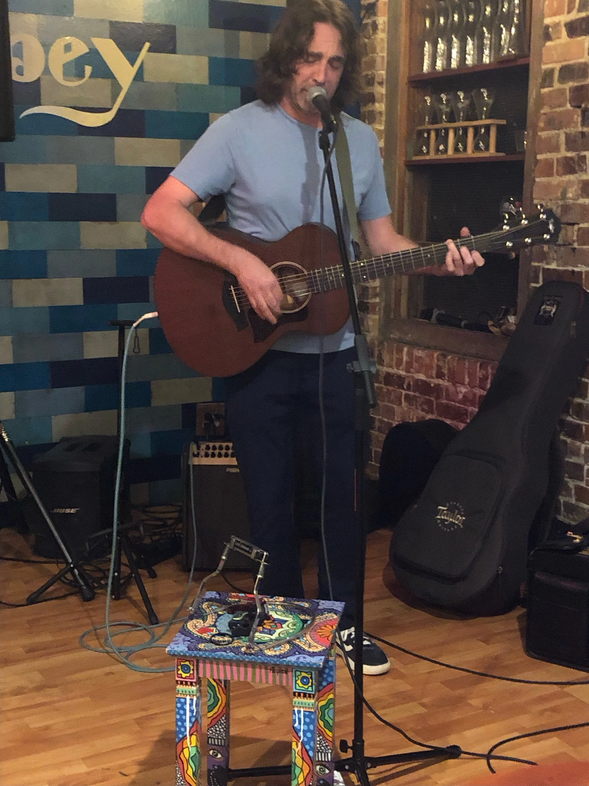 Man playing acoustic guitar and singing into a microphone at a live music venue with brick walls and colorful decor.