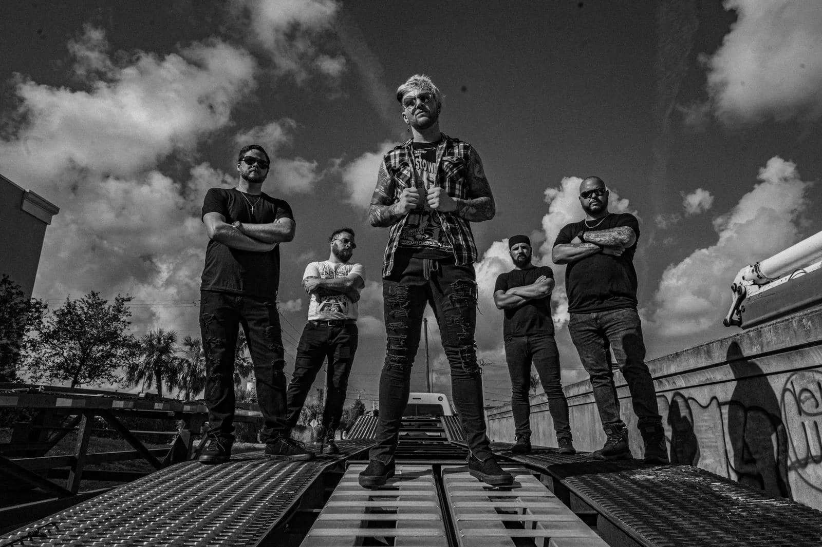 Black and white photo of six men standing on a rooftop, with crossed arms, looking at the camera. There are clouds in the sky and graffiti on the wall behind them.