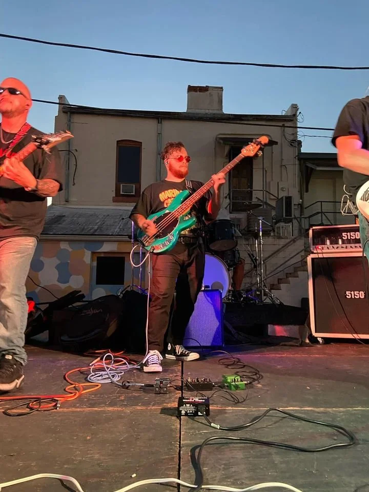 Musicians performing on an outdoor stage at dusk, playing guitars and bass, with various equipment and amplifiers visible.