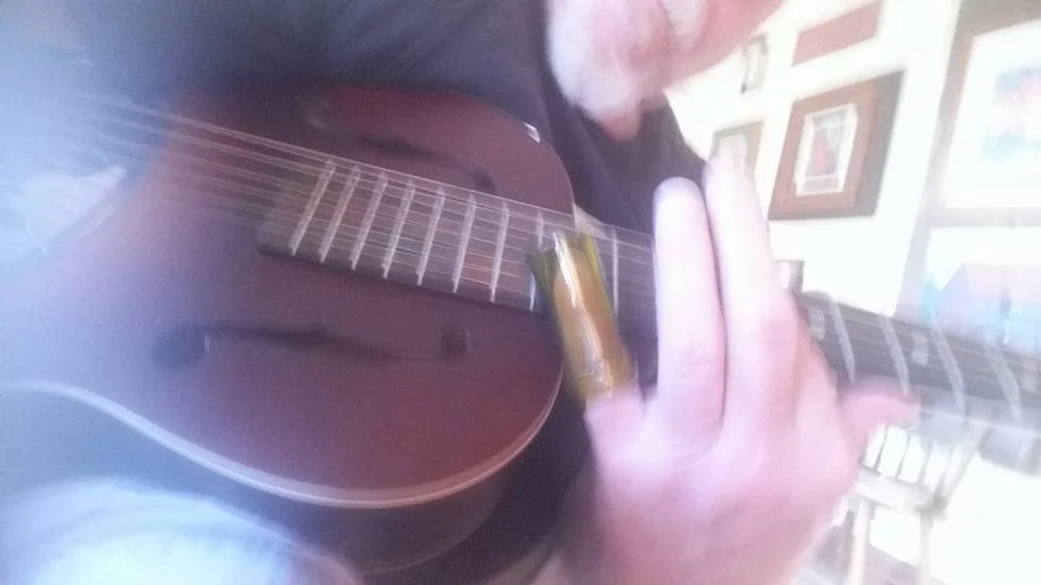 Kim Kimbo Groff playing a guitar, close-up on hands and guitar body, with a background of framed pictures on a wall.