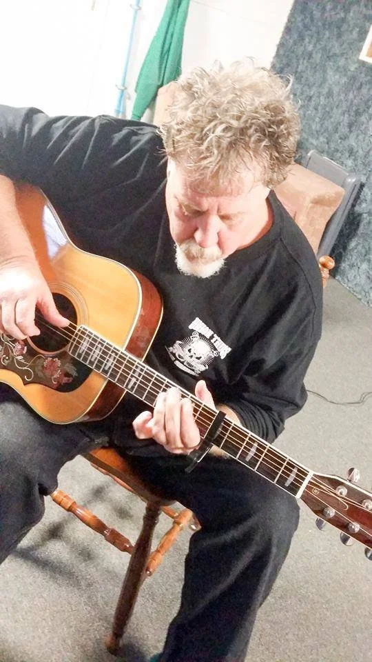 Kim Kimbo Groff with curly gray hair and a beard, wearing a black shirt, sitting on a wooden chair and playing an acoustic guitar.