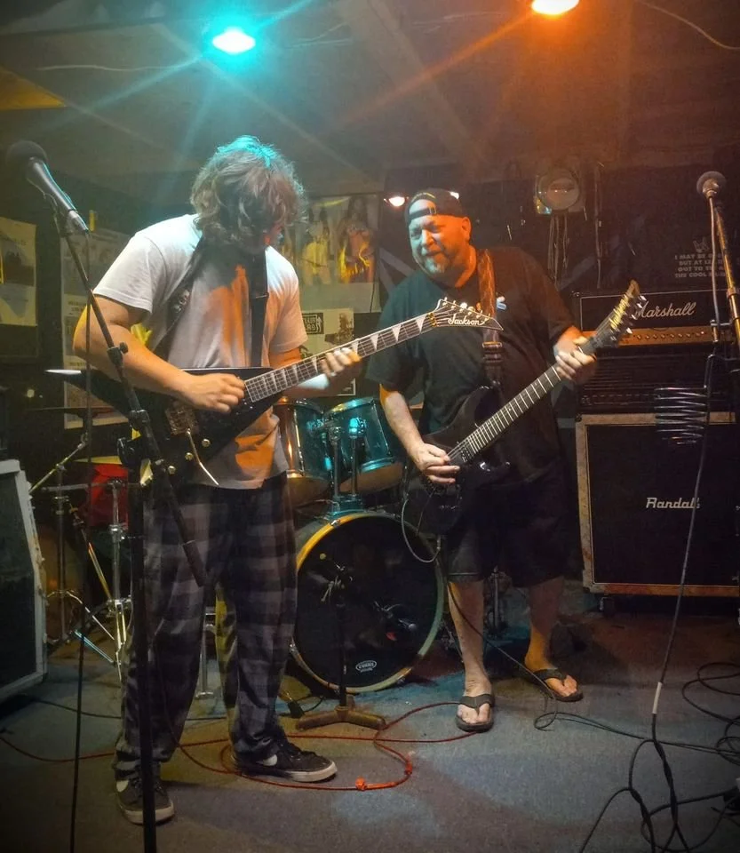 Two men playing electric guitars on stage in a dimly lit venue with colorful stage lights, a drum set behind them, and Marshall amplifiers on the right.