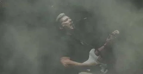 A musician playing an electric guitar amidst fog or smoke with a joyful expression.