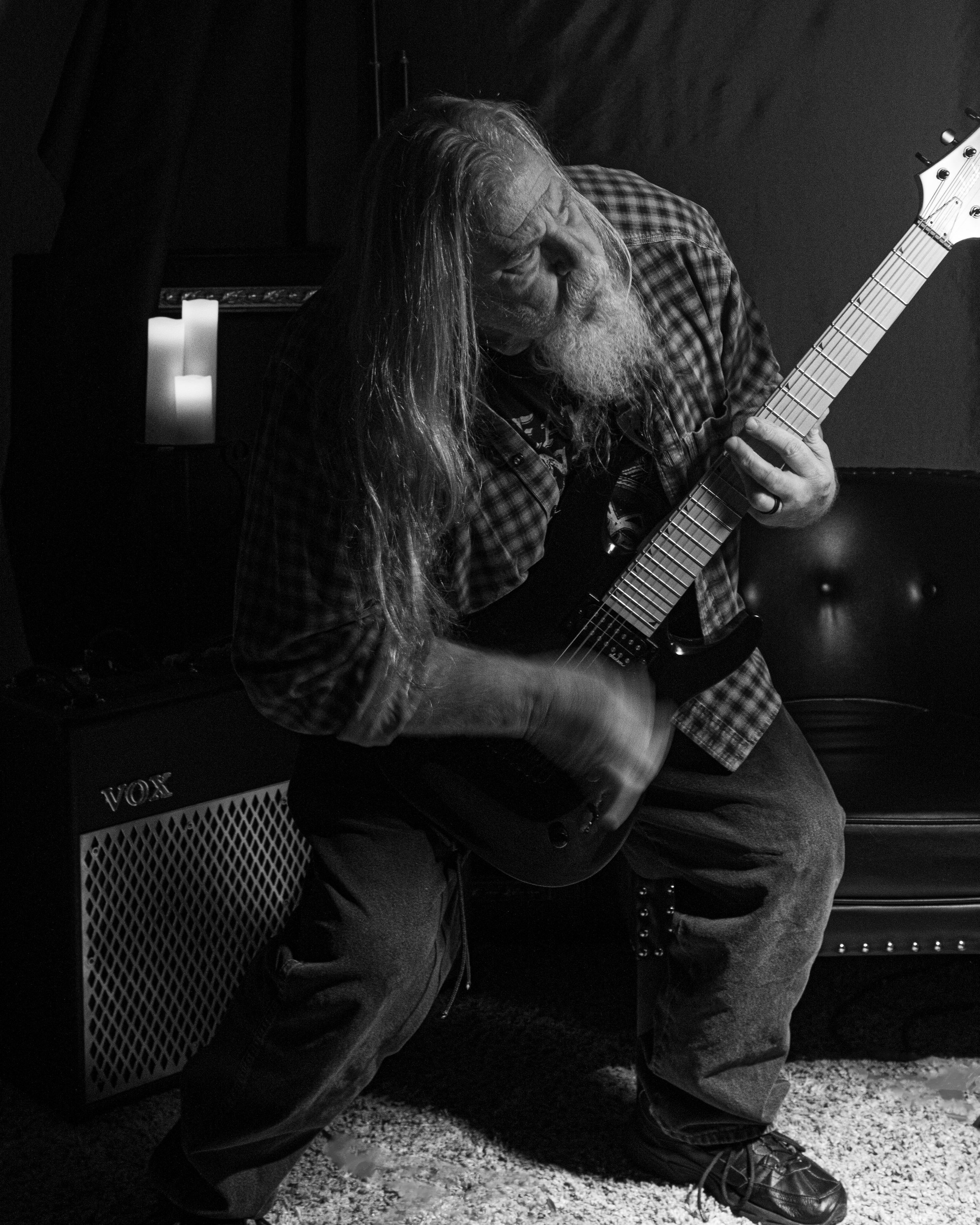 A photo of Kim Kimbo Groff with long hair and a beard playing an electric guitar in a dimly lit room, sitting on a bench next to a small amplifier and a lit candle.