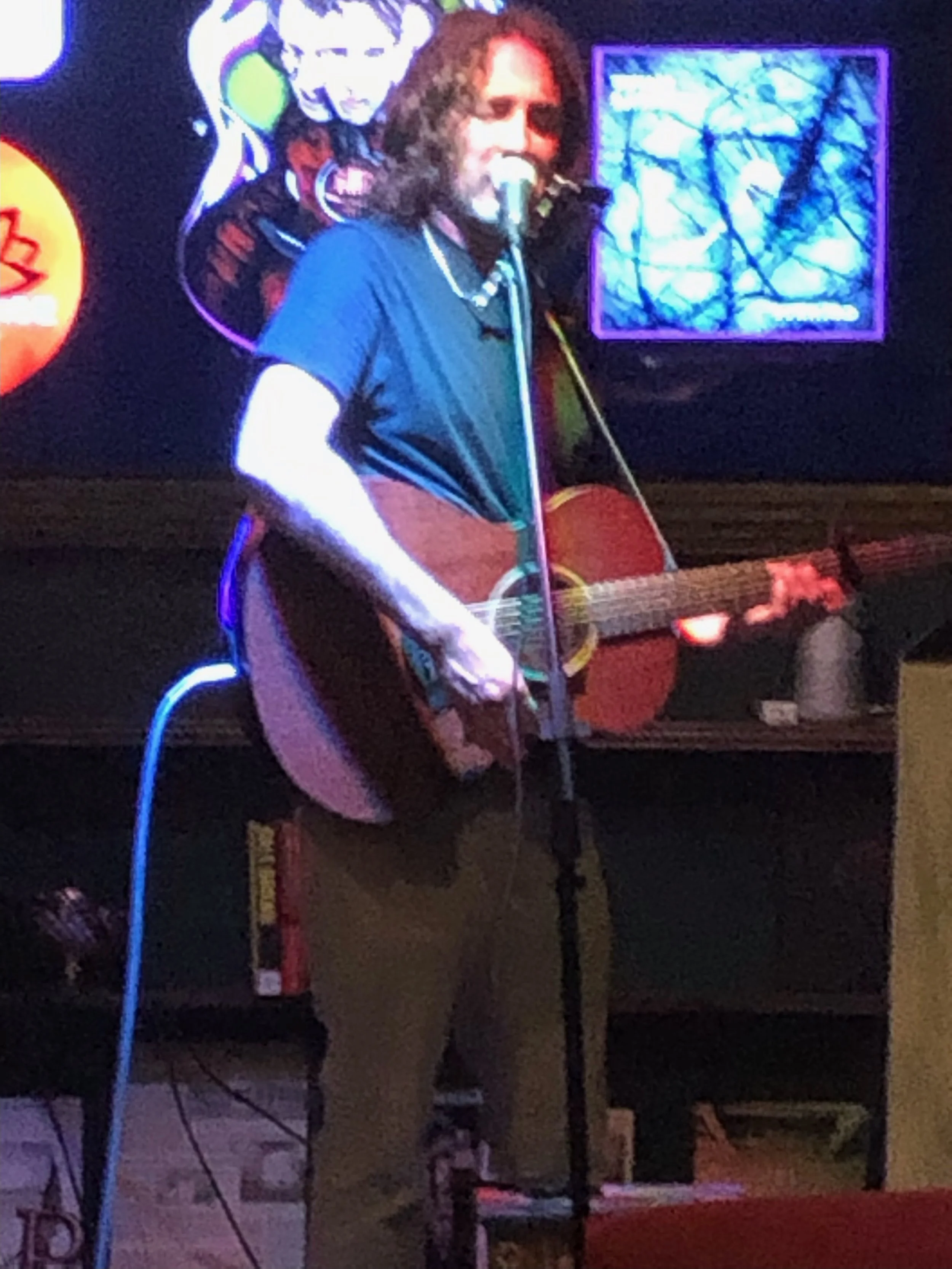 A man with curly hair and a beard playing an acoustic guitar and singing into a microphone on a stage with neon artwork in the background.