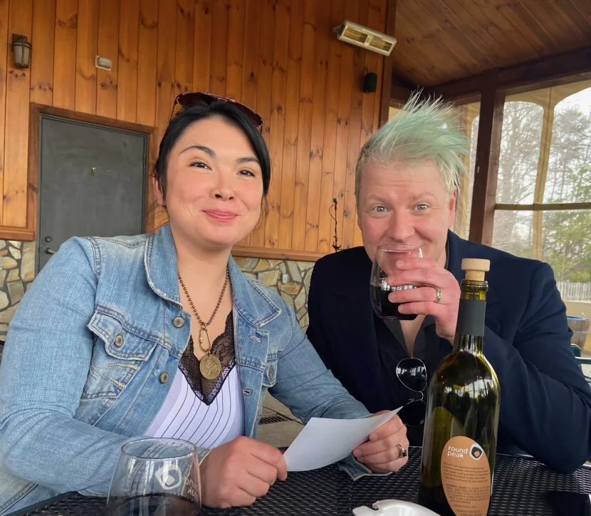 A woman and a man with light green spiked hair sitting at a table in a wooden cabin, smiling and drinking from a glass of red wine. There is a bottle of wine and a glass on the table.