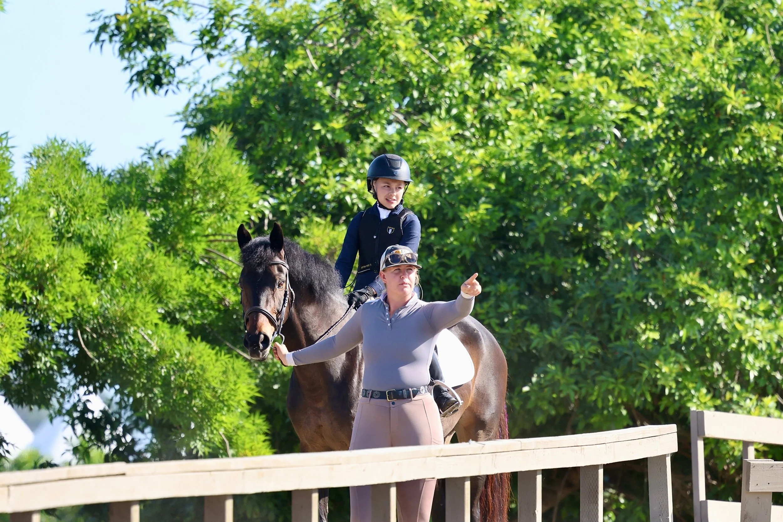 A young beginner rider sits confidently on a dark horse, guided by an instructor pointing ahead during a horseback riding lesson at Strides Riding Academy.
