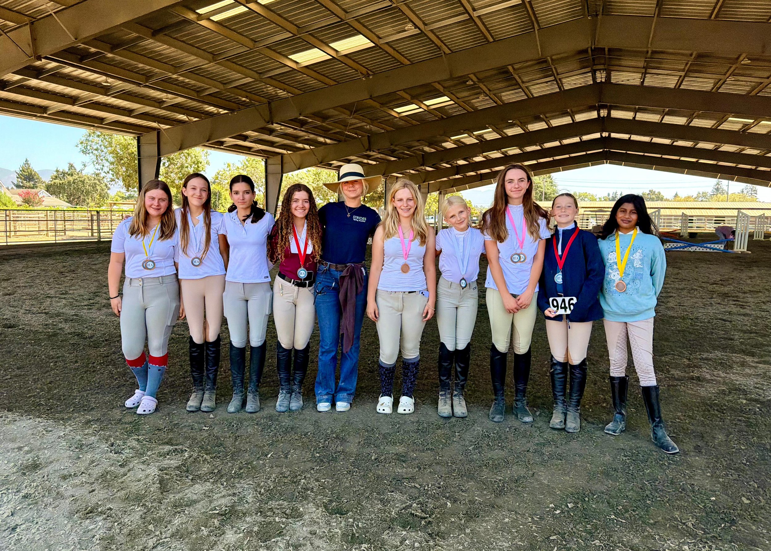 Group photo of the Strides IEA riding team and trainer posing together inside a covered arena with their show ribbons around their necks after their first competition.