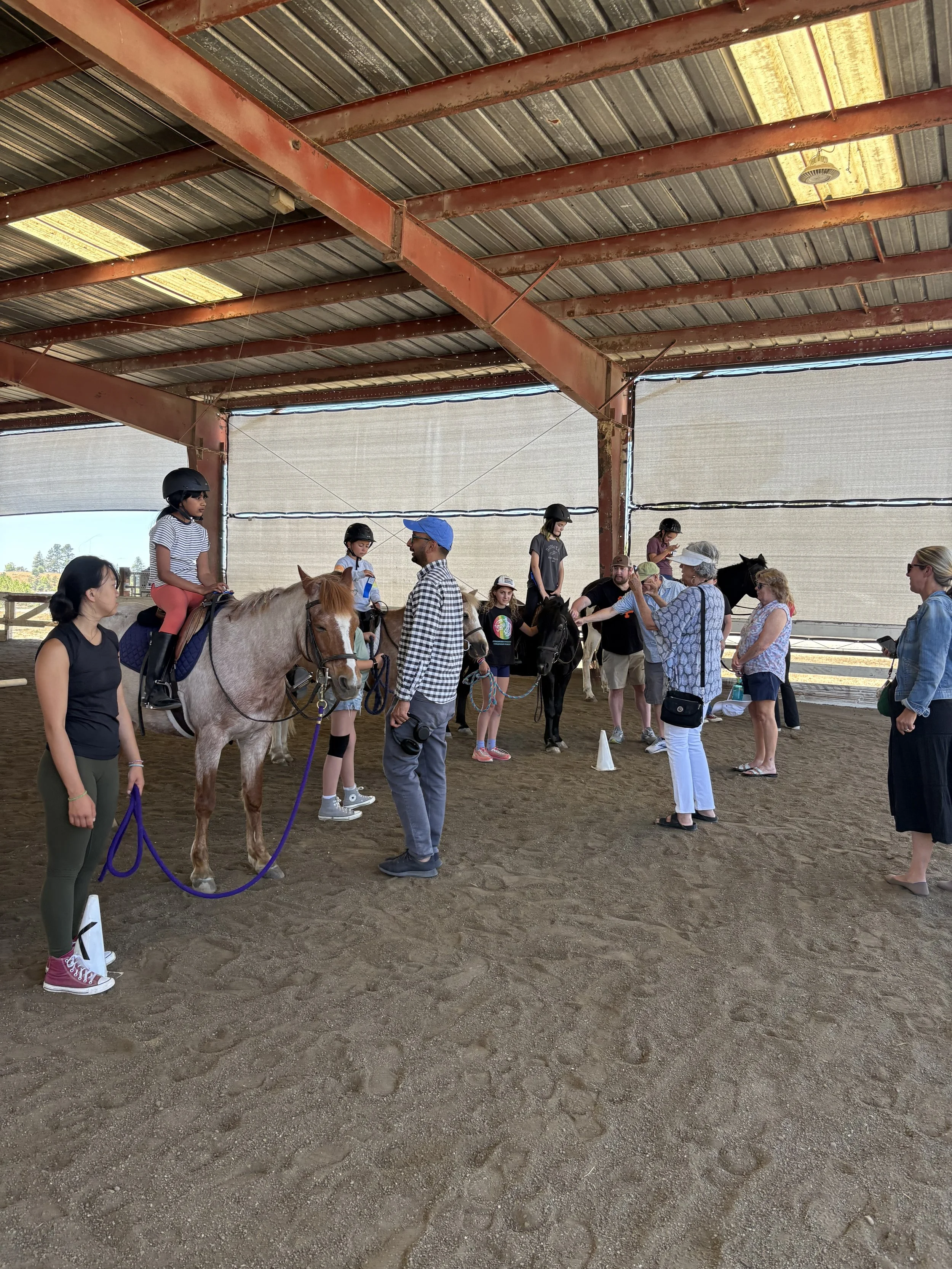 Children participating in a summer horseback riding camp at Strides Riding Academy in Sonoma County, riding horses with instructors while parents observe in a covered arena.