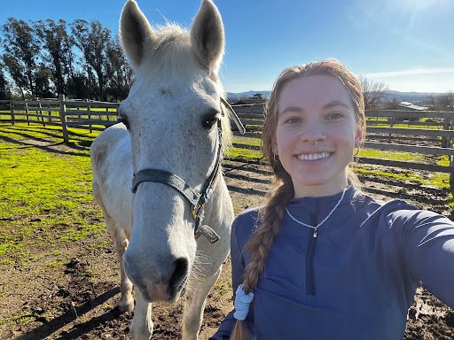 Adult horseback rider smiling beside a white horse at Strides Riding Academy in Petaluma, California, on a sunny summer day.
