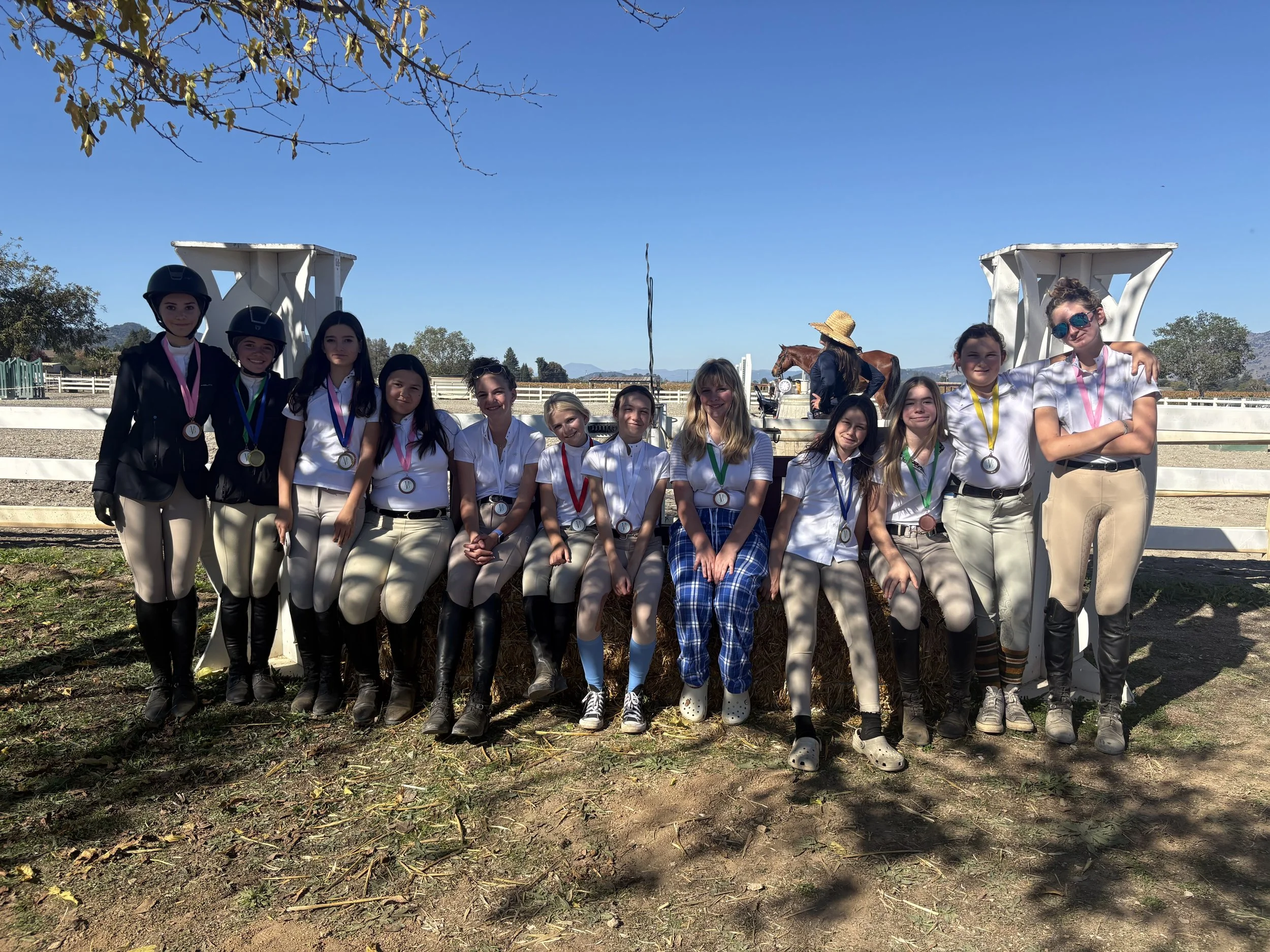 A group of young riders from Strides Riding Academy posing together outdoors after a November horse show, wearing medals and show attire, with horses and the arena in the background.