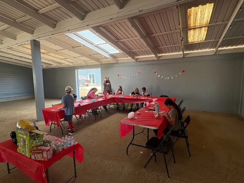 Children and families participating in school-themed crafts around red-covered tables set up inside a covered barn space during the Back-to-School Barn Bash.