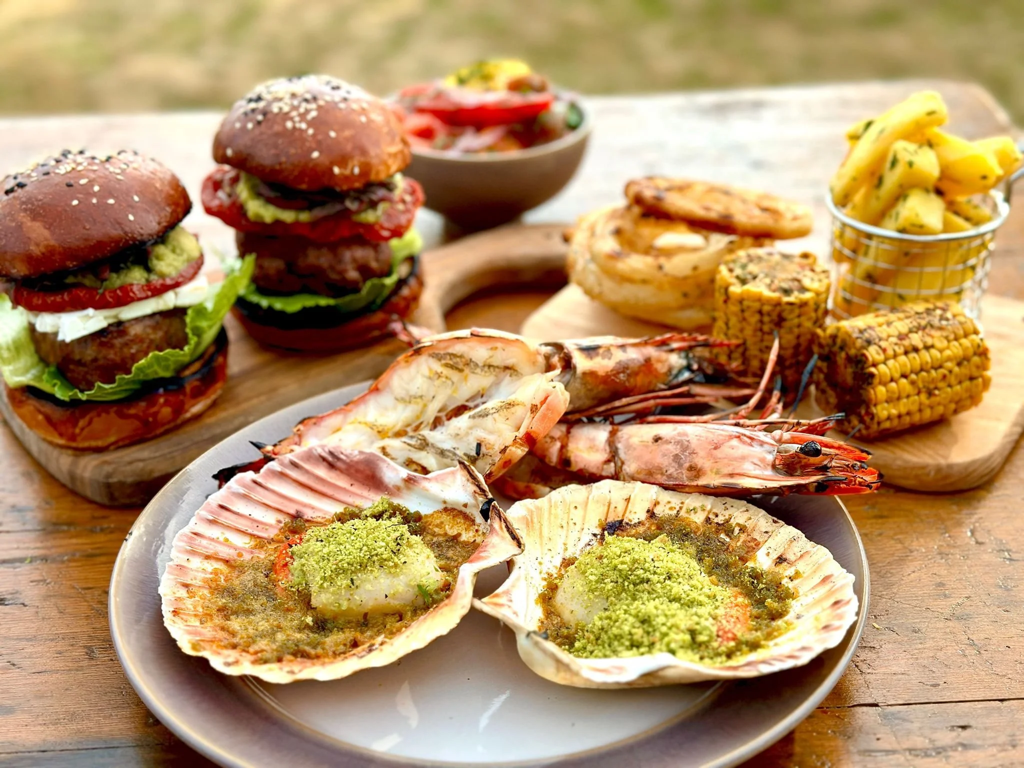 Seafood including baked scallops with green seasoning, grilled prawns, and grilled corn on the cob, with burgers and salads in the background.